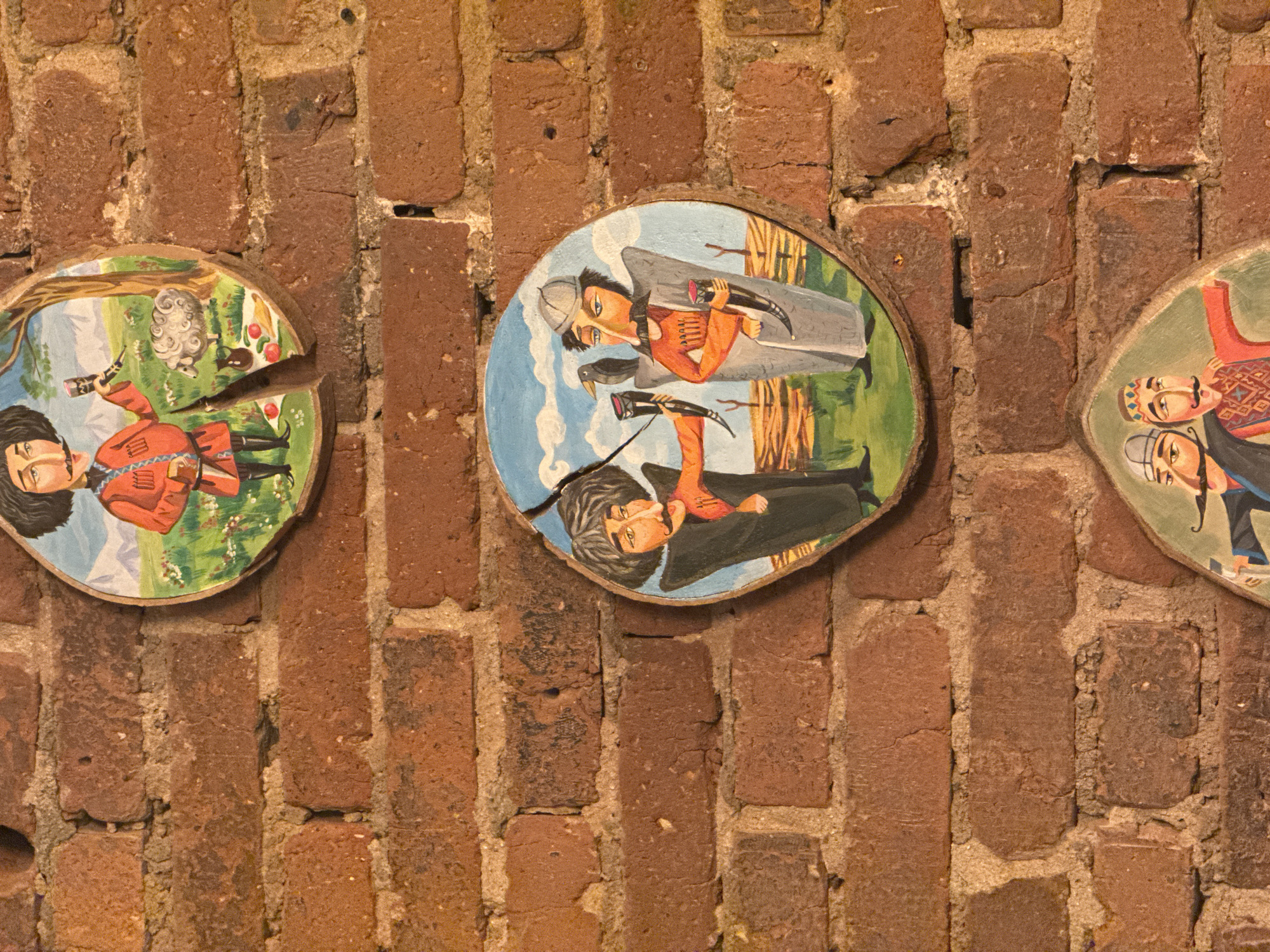 Three circular, folk art paintings depicting men in traditional attire are displayed on a brick wall. The top painting shows a man in a red tunic and hat holding a goblet, with a sheep and a mountainous landscape in the background. This painting appears to be broken into two pieces. The middle painting features two men, one in a red tunic and hat, the other in a grey outfit and helmet, both holding drinking horns. A crow perches on the shoulder of the man in the grey outfit. The bottom painting, partially visible, shows two men with mustaches and traditional headwear. The paintings are mounted on a rustic red brick wall, which shows signs of age and some damage. The lighting suggests an indoor setting, possibly a restaurant or cafe. The location of these paintings in Chelyabinsk, Russia, is unknown.