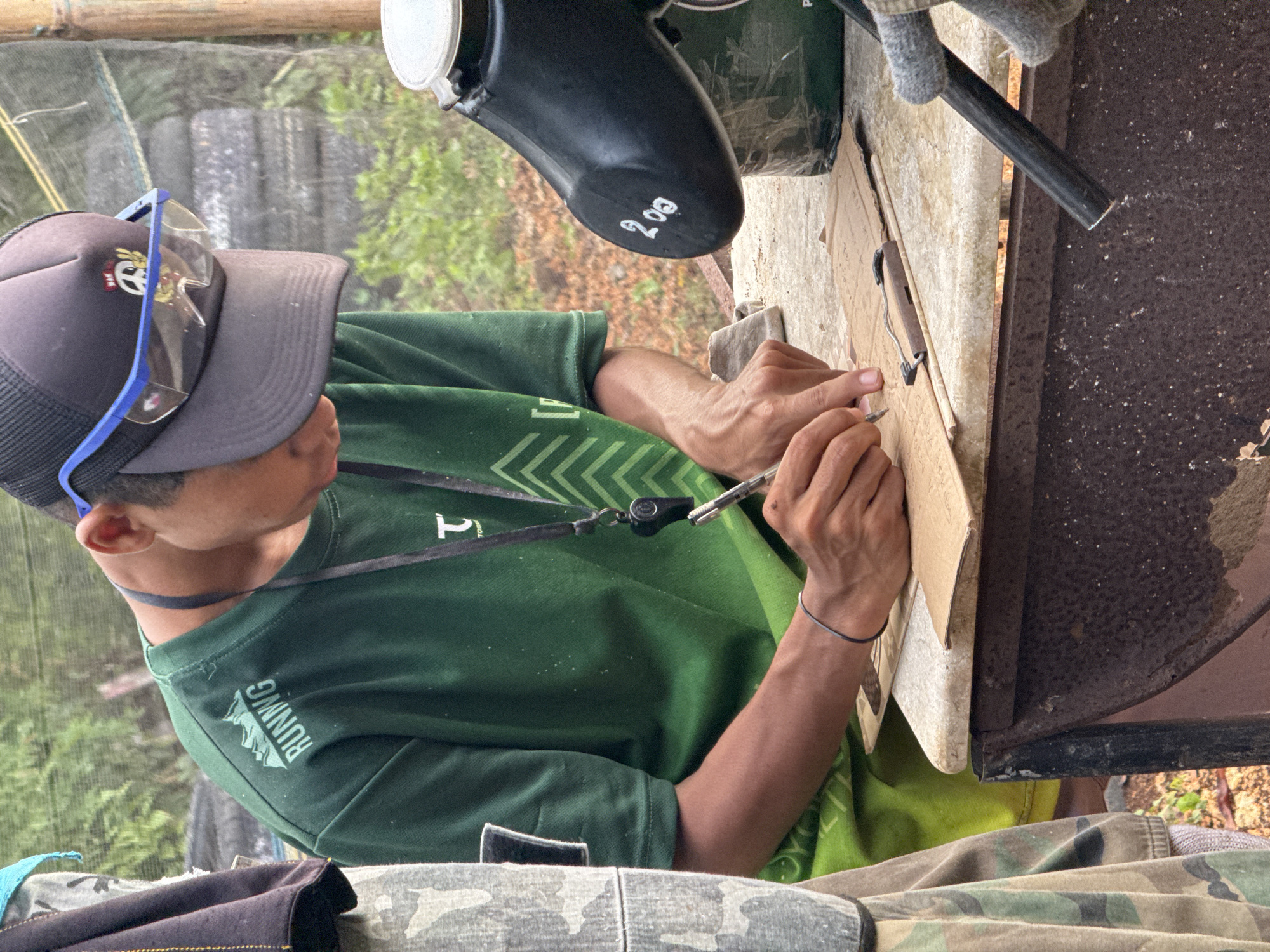 A person is working with a cardboard box and a tool resembling a box cutter on top of a surface made of weathered materials. The person wears a grey cap with a logo, safety glasses, a green t-shirt with the word "RUNNING" visible, and a black strap around their neck. Camouflage-patterned clothing is visible below the table surface. Above the table is a black object with the number "208" written on it. The location is Kuta Selatan, Indonesia.