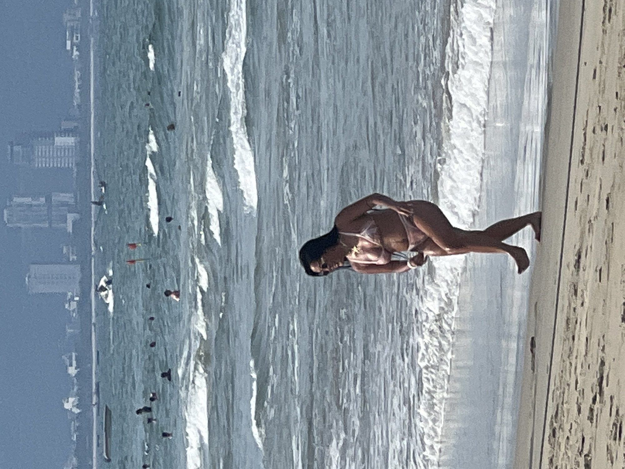 The image shows a woman in a pink bikini walking on a sandy beach with the ocean and city skyline in the background. The waves are breaking near the shore. In the distance, several boats and jet skis are visible on the water, and a hazy city skyline with tall buildings can be seen. The sky is overcast. The woman is facing to her left, with her head turned to her right, and she is smiling. She has sunglasses on and is wearing a necklace. Her right arm is bent at the elbow, and her left arm is extended slightly behind her. Her legs are positioned as if she is walking or dancing. The beach has footprints in the sand. The overall scene appears to be a summer day at a beach resort area in Đà Nẵng, Vietnam.