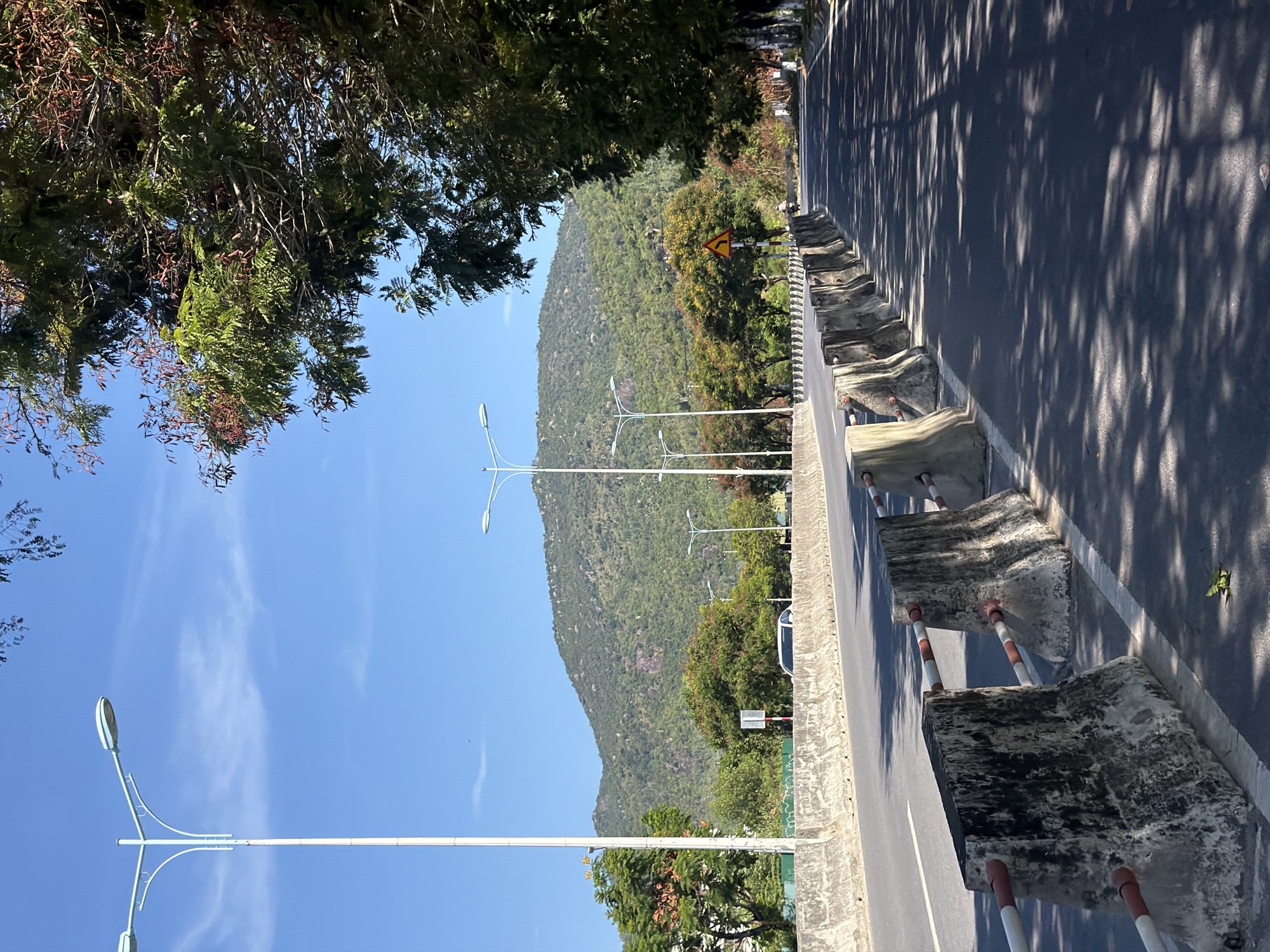 The image shows a street scene in Nam Nha Trang Ward, Vietnam. In the foreground, a road is lined with concrete barriers that are weathered and stained. The road itself is asphalt with white lane markings. Sunlight filters through trees on the right side of the road, casting dappled shadows on the pavement. In the distance, a large, tree-covered hill dominates the landscape under a clear blue sky. Several streetlights line the road, and a white car is visible on the opposite side of the divided highway. There are some trees and vegetation alongside the road, with a few small red-brown seed pods on some of the trees. A single person on a motorcycle is visible on the road in the distance.