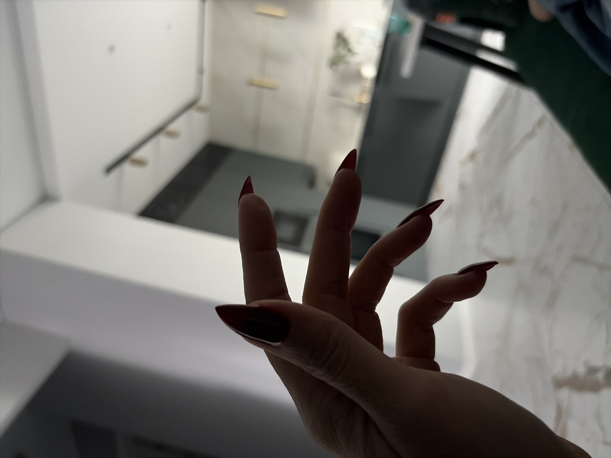 The image shows a close-up of a hand with long, pointed, dark red fingernails. The hand is positioned in the foreground, obscuring the view of what is behind it. In the background, a modern kitchen or living area is visible, featuring dark cabinetry, a light-colored countertop, and some decor items. There are also pendant lights hanging from the ceiling. The flooring appears to be light-colored marble or tile. The overall lighting suggests an indoor environment, possibly in the evening or with dim ambient light. There is no discernible information about the location being Moscow, Russia.