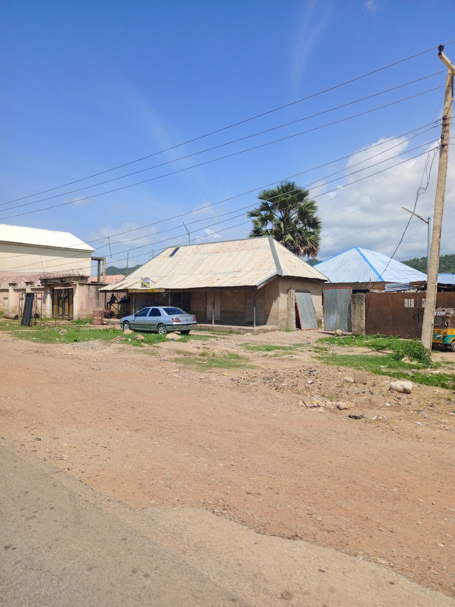 The image depicts a street scene in Tilde, Nigeria, under a clear blue sky with scattered clouds. In the foreground, a dirt road transitions to a paved surface. On the right side of the road, a concrete utility pole with multiple wires and a street lamp stands tall. Further into the scene, various structures line the road.
To the left, a light-colored, multi-story building with a white roof is partially visible, featuring a fence with barbed wire. Adjacent to it, a single-story building with a corrugated metal roof and stone block walls is present. A silver sedan is parked in front of this building.
In the center of the image, a low building with a corrugated metal roof displays a sign reading "A-B Gas". Two individuals are seated outside this establishment, and a few barrels are visible nearby. Behind this building, a large palm tree with distinctive fronds stands out.
Further to the right, another building with a blue, corrugated metal roof is situated behind a brown metal fence. A small, yellow, three-wheeled vehicle, possibly a tuk-tuk, is parked near the fence, displaying a license plate that appears to read "4-1186". The ground surrounding the buildings is covered with sparse green grass and patches of dirt, with some scattered debris. The overall impression is that of a typical roadside scene in a Nigerian town.