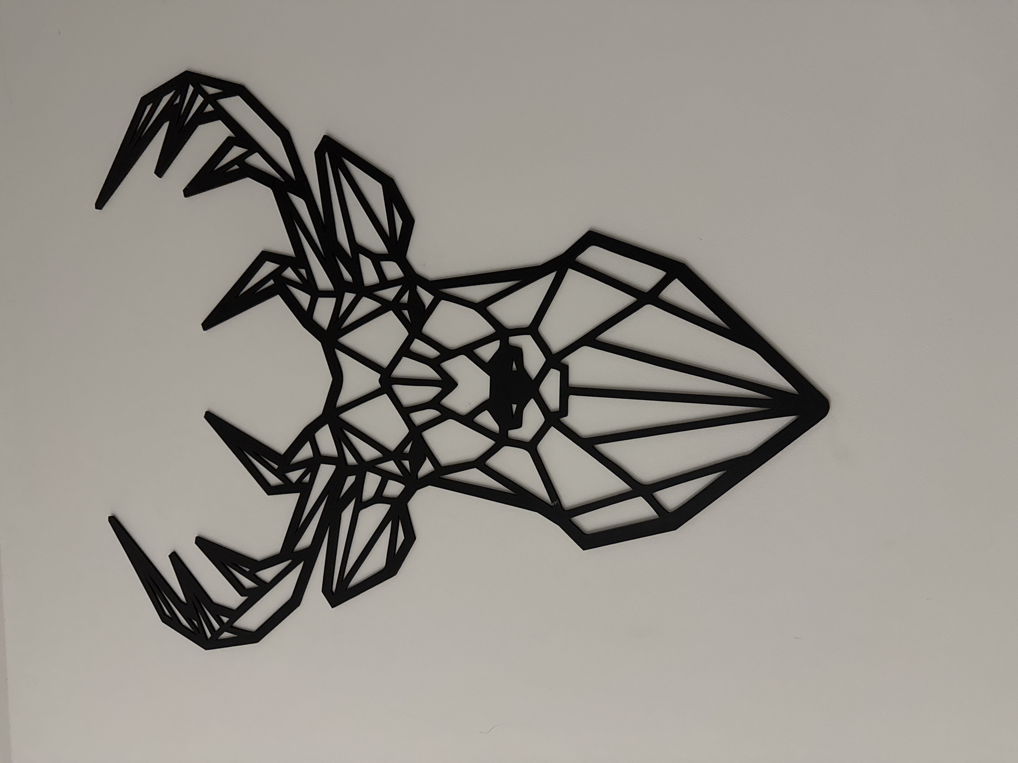 A black, geometrically constructed deer head silhouette is centrally mounted on a plain, light-colored wall. The decoration depicts the head and prominent antlers of a deer, rendered entirely through a network of interconnected straight lines forming various triangular and polygonal shapes. The antlers extend upwards and outwards from the head, exhibiting multiple points and branches, each formed by distinct geometric segments. The head itself is composed of angular forms defining the cheeks, snout, and an implied nasal area at the bottom center. The object appears to be a flat, solid material, possibly metal or wood, with uniform thickness and clean edges, casting minimal shadows against the background, suggesting even illumination. No other objects or individuals are visible in the scene.