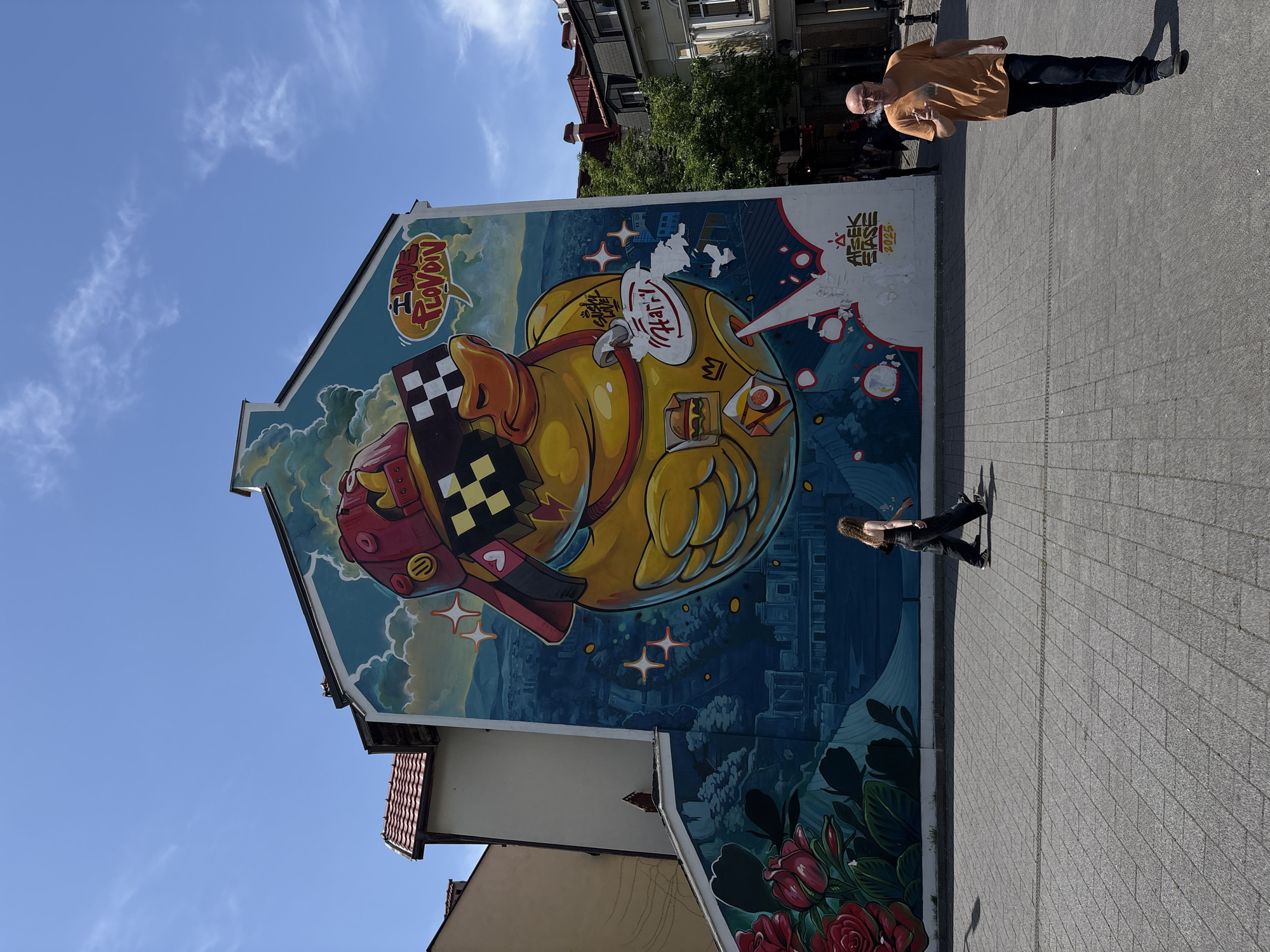 A vibrant mural covers the side of a building in Plovdiv, Bulgaria. The central image is a large yellow cartoon duck wearing pixelated sunglasses and a red rocket-shaped hat with black and white checkered designs. The duck has a speech bubble saying “Yummy” next to it. Other design elements include cartoon food items, stars, flowers, and a city-like landscape in the background. Two people are walking on the sidewalk next to the building and mural. The sky is clear and blue.