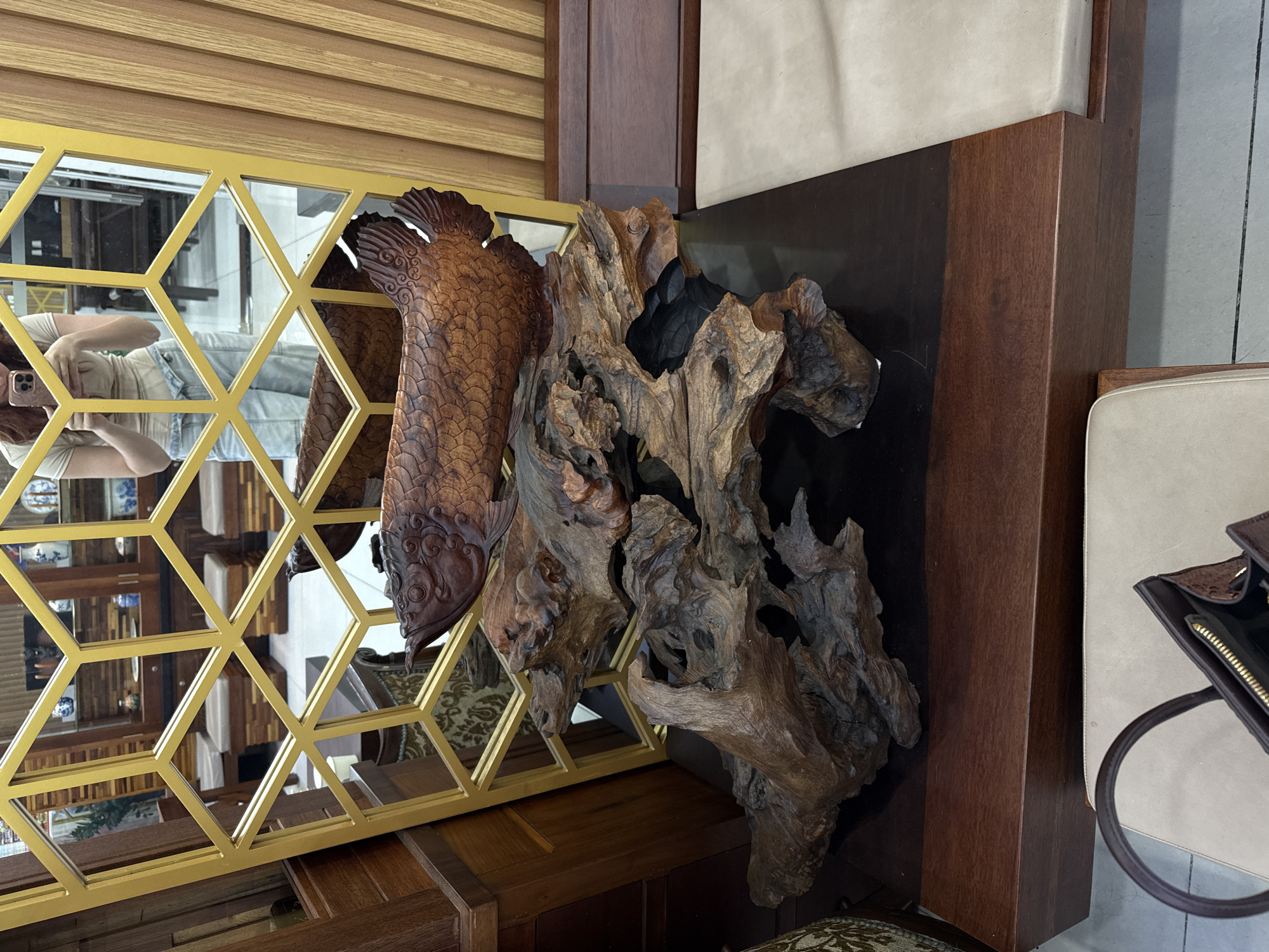 This indoor scene captures a wooden carving of a fish beside a piece of driftwood, both displayed in front of a geometrically patterned mirror. The mirror reflects the surroundings, including the individual holding the camera. The location is Tay Nha Trang Ward, Vietnam.