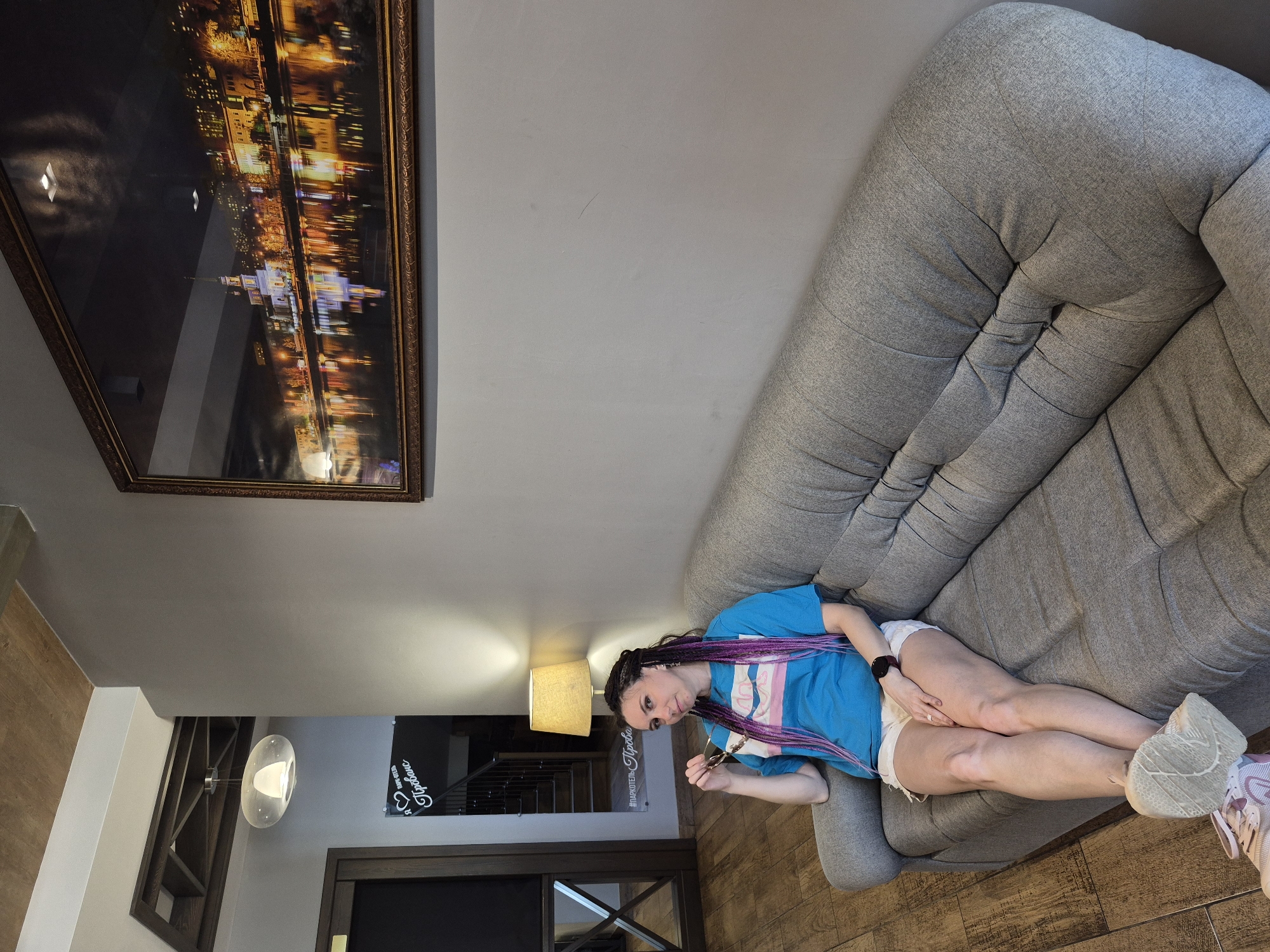 A woman is lying on a gray couch, wearing a blue t-shirt, white shorts, and sneakers. Her hair is styled in braids with purple highlights. She is holding sunglasses. The room contains a framed picture hanging on the wall, a lamp, and a door with a sign written in Russian. The sign includes the word "votkinskie", possibly indicating the location in Votkinsk, Russia. The floor is wooden.
