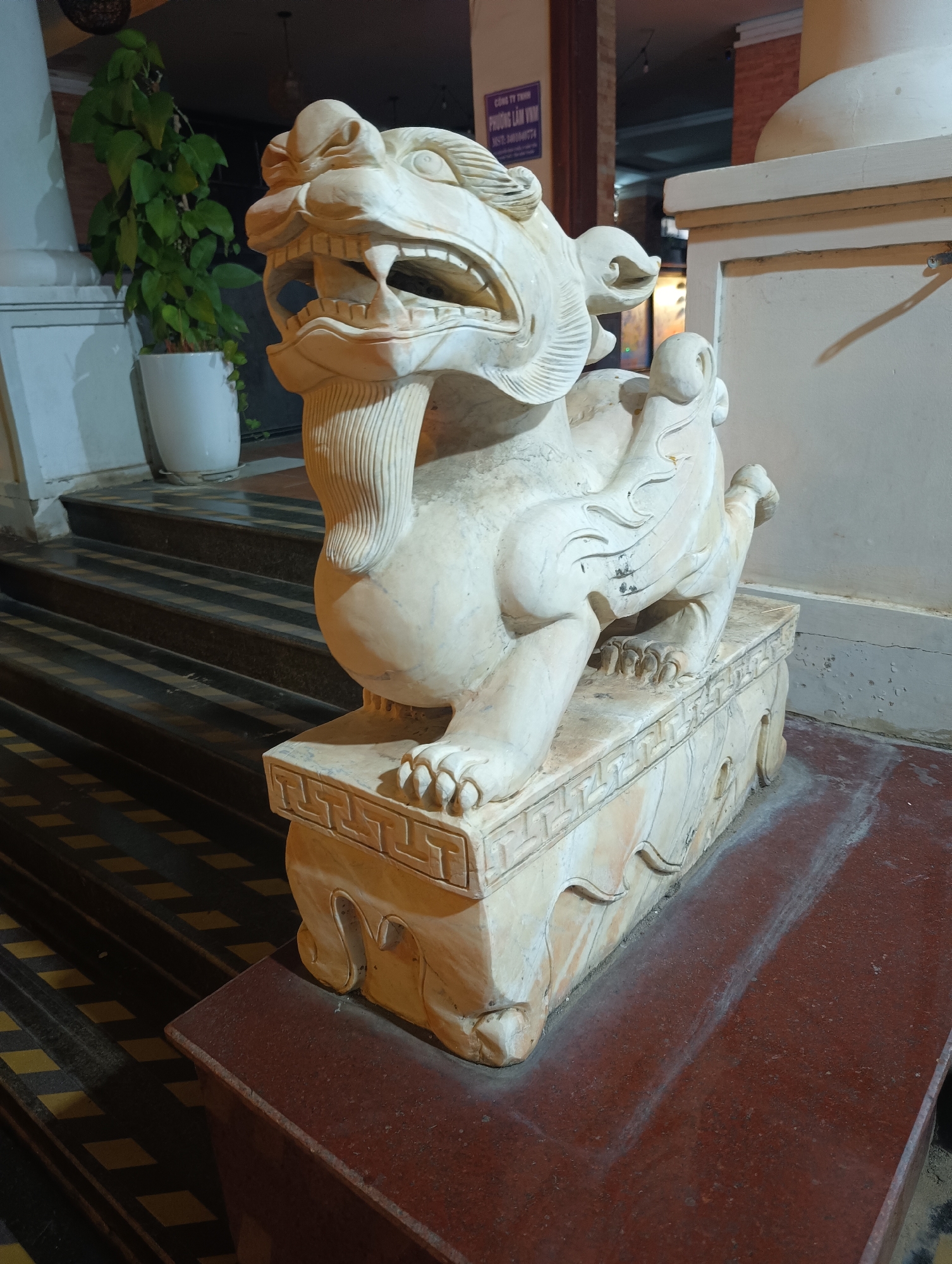 A white marble sculpture of a mythical creature, possibly a Foo dog or lion, is positioned on a reddish-brown granite base in front of a building at night. The sculpture is detailed, with an open mouth, sharp teeth, and flowing mane. It sits on a rectangular pedestal adorned with geometric patterns and carved details. Behind the sculpture, a set of dark stairs with yellow and black striped markings leads up to an entrance. A potted green plant stands to the left of the stairs. The building's facade features a sign with text that appears to be in Vietnamese, and some brickwork is visible. The lighting suggests an outdoor or semi-outdoor setting illuminated by artificial light sources. The location is identified as Phu Thuy Ward, Vietnam.
