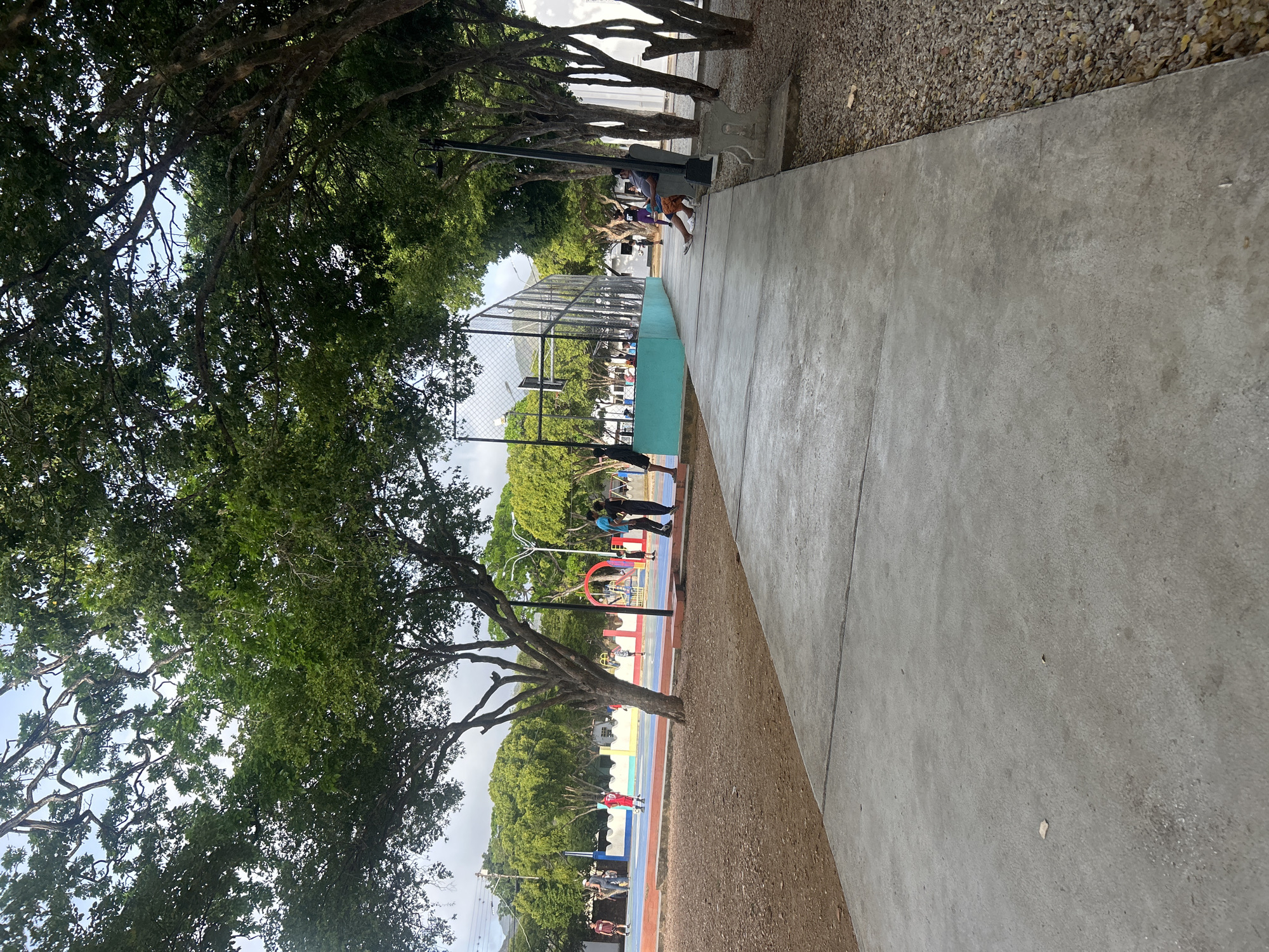 The image shows an outdoor public space with a concrete walkway and a dirt area. In the background, there is a sports court with a fence and several people engaged in various activities. Some individuals are playing on the court, while others are observing. Lush green trees with dense foliage line the walkway and the sports court, providing shade. The sky is partly cloudy. The scene suggests a community gathering space or recreational area within the city of Santa Ana, Venezuela.