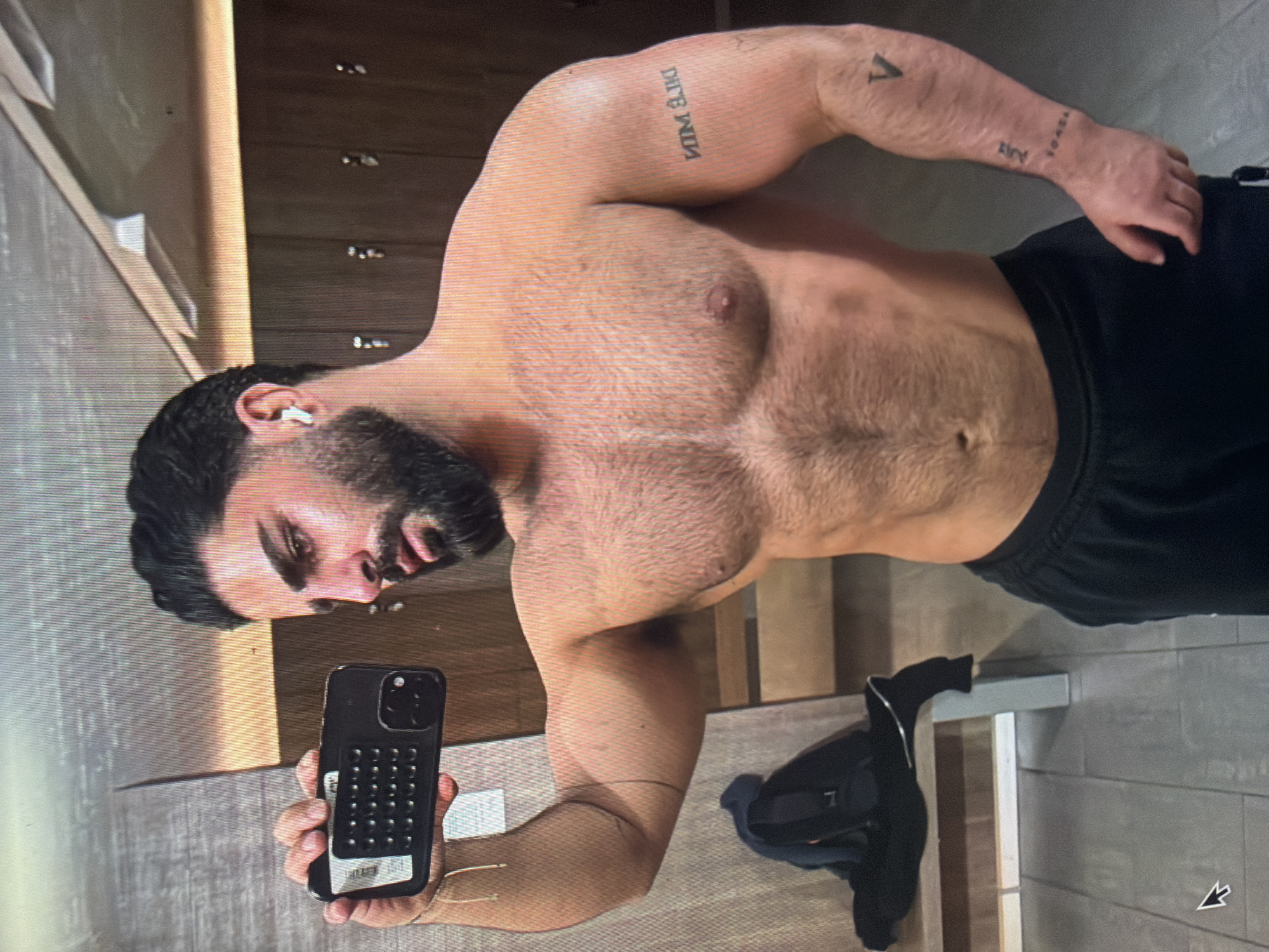 A muscular adult male with dark hair, a beard, and a mustache is depicted shirtless, wearing black athletic shorts, and a white earbud in his right ear. He is taking a mirror selfie, holding a black smartphone with a textured case in his left hand. Visible tattoos include mirrored text on his left bicep, a 'V' symbol on his right forearm, and an inscription on his right wrist/forearm. The scene is an indoor locker room or changing facility located in Shumen, Bulgaria, featuring dark wood-paneled lockers, strip lighting, a light grey tiled floor, and a light-colored bench with a dark bag or garment resting on it. A mouse cursor is present in the lower-left corner of the overall image.