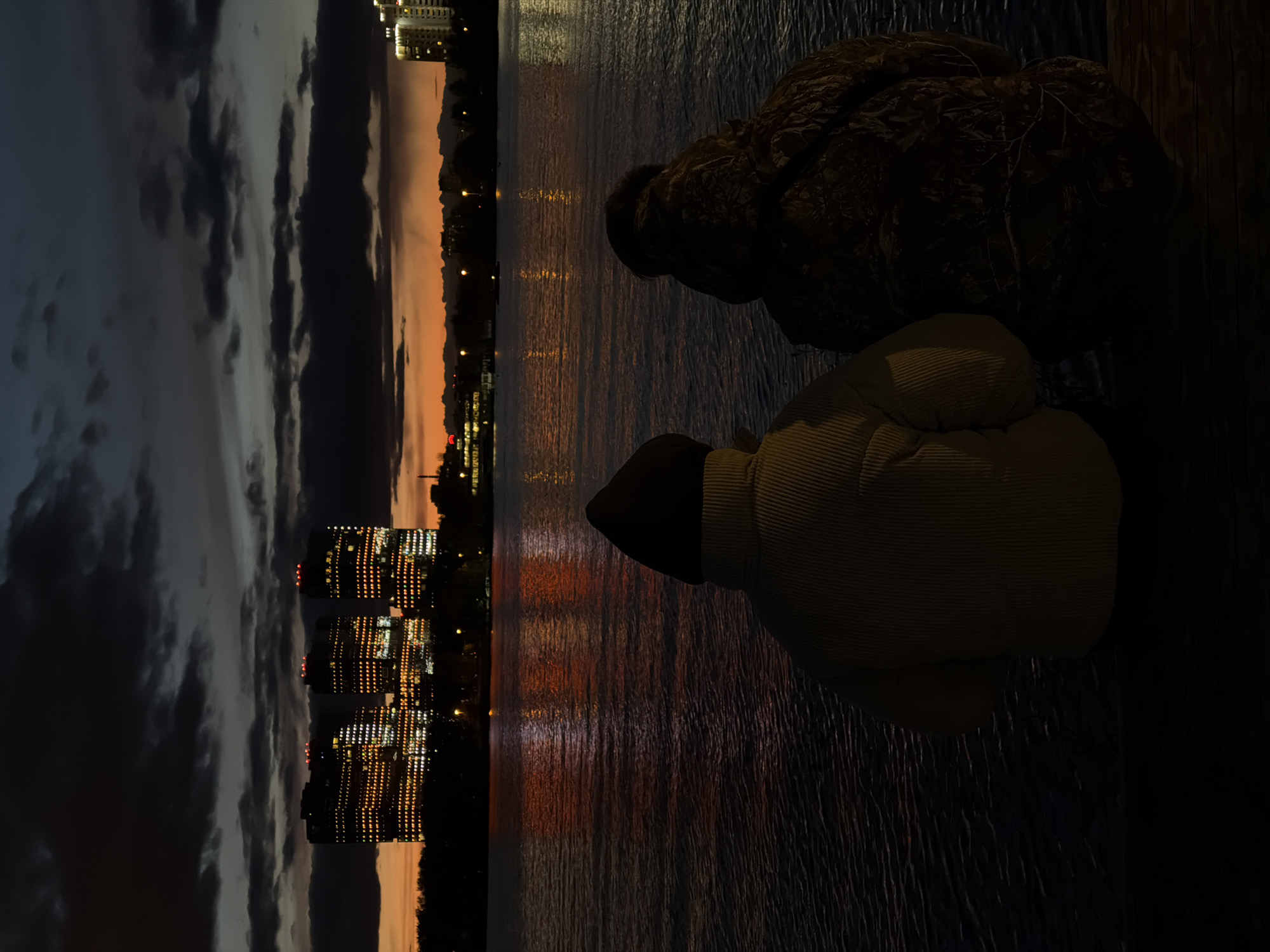 The image shows a nighttime view in Kazan, Russia, likely taken from the bank of the Kazanka River. In the distance, illuminated high-rise buildings reflect in the dark water, their lights scattered across the surface. The sky above displays a dramatic mix of dark clouds and a band of orange light near the horizon. Closer to the viewer, two figures are silhouetted against the scene. One appears to be a person wearing a hooded jacket, seated with their back to the camera. Next to them is a dark, rounded object, possibly a rock or another person. The foreground consists of what seems to be a dark, textured wooden surface, perhaps a deck or a walkway along the riverbank.
