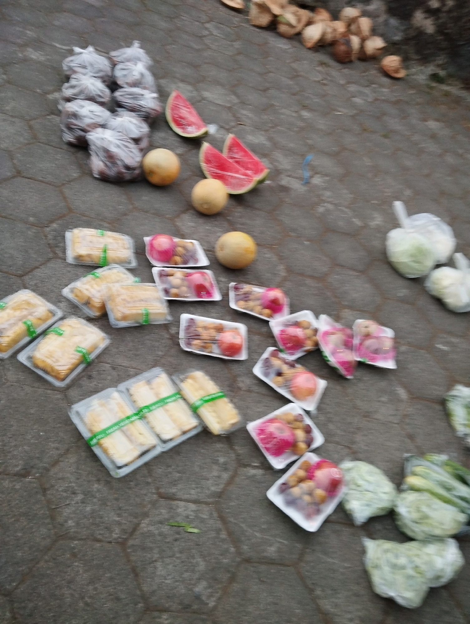 The image shows a collection of food items, including fruit and vegetables, arranged on a paved surface that appears to be a sidewalk or street in Salatiga, Indonesia. The food is packaged in plastic and some is unwrapped. There are several plastic-wrapped bundles of what look like root vegetables or small fruits, along with sliced watermelons, and containers of mixed fruits. There are also whole fruits like melons and cabbages, some of which are wrapped in plastic. In the background, there are pieces of dried coconut shells. The scene suggests a market stall or a roadside vendor's display. The lighting is overcast, and the pavement is a dark gray hexagonal tile.