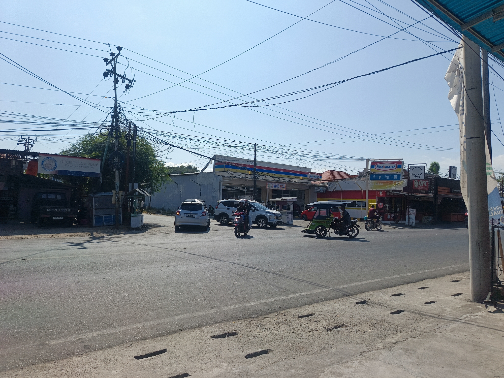 The image depicts a street scene in Luwuk, Indonesia, on a clear day under a bright blue sky. The foreground shows a paved road with visible traffic markings and a concrete sidewalk with drainage grates. In the mid-ground, various vehicles are present, including a silver sedan with license plate BM 1473 GA, a white SUV, a motorcycle with a rider, and a three-wheeled vehicle, often referred to as a "bajaj," carrying a passenger. A red vehicle is parked in the background.
Several commercial establishments are visible along the roadside. A prominent Indomaret convenience store with its characteristic red, yellow, and blue signage is featured. Other businesses with various signs, some in Indonesian, are also present, including one advertising "Semen Tiga Roda" (Three Wheel Brand Cement). A tall utility pole with a complex network of tangled electrical wires dominates the left side of the frame, casting shadows on the road. A dark-colored Mitsubishi truck is parked near the utility pole. The environment appears to be an urban or semi-urban area.