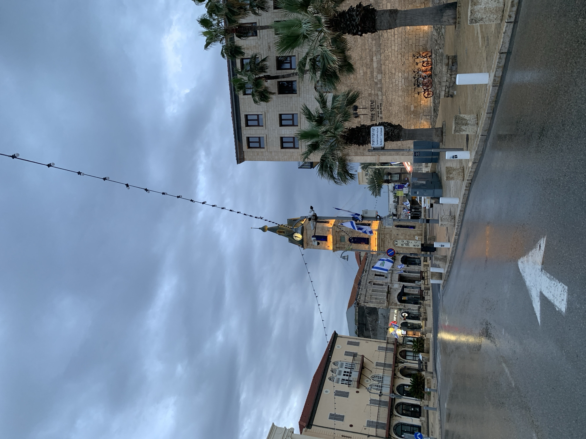 The image depicts a street scene in Tel Aviv, Israel, under an overcast sky. The foreground features a wet road with a white arrow indicating the direction of traffic. The clock tower of Jaffa is visible and is decorated with Israeli flags. A string of lights spans the street. Adjacent to the clock tower are buildings with arches and balconies. The text "RIP CURL" is visible on one of the buildings. Palm trees and additional buildings, including "THE SETAI", are also in the background.