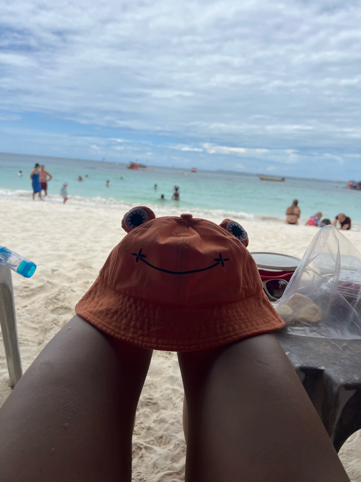 The image is a first-person perspective shot taken at a beach in Pattaya City, Thailand. The foreground is dominated by the photographer's legs, with an orange bucket hat featuring a frog design resting on their lap. To the left, a snorkel and mask are visible, partially obscured by a chair. On the right, a plastic bag containing what appears to be food items sits on a dark surface, likely a table. In the mid-ground, the white sandy beach is populated by several people, some wading in the shallow turquoise water, while others are seated on the sand. The background shows the calm sea under a partly cloudy sky. Boats are scattered in the distance on the water. The overall impression is a relaxed day at the beach in Pattaya.
