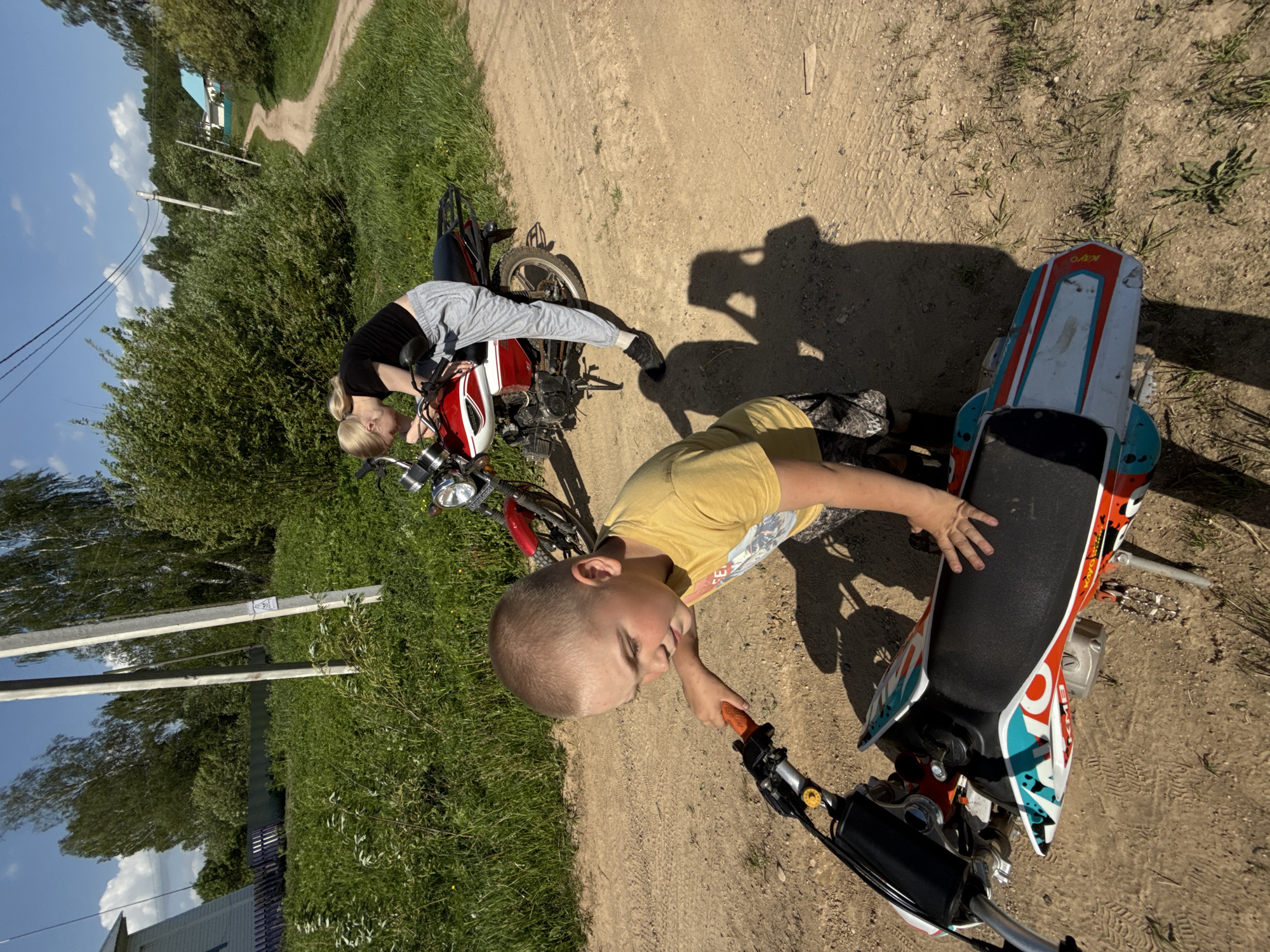 The image shows two people with motorbikes on a dirt road. A child in a yellow shirt is on a small dirt bike with red, white, and blue decorations. An adult female is leaning over a red and white motorbike. The scene is located in Zhigalki, Russia, as indicated by the location metadata. The environment consists of a dirt road, grass, trees, and a few buildings in the distance. Shadows cast by the subjects suggest it is daytime. The road stretches into the distance, winding through the landscape.