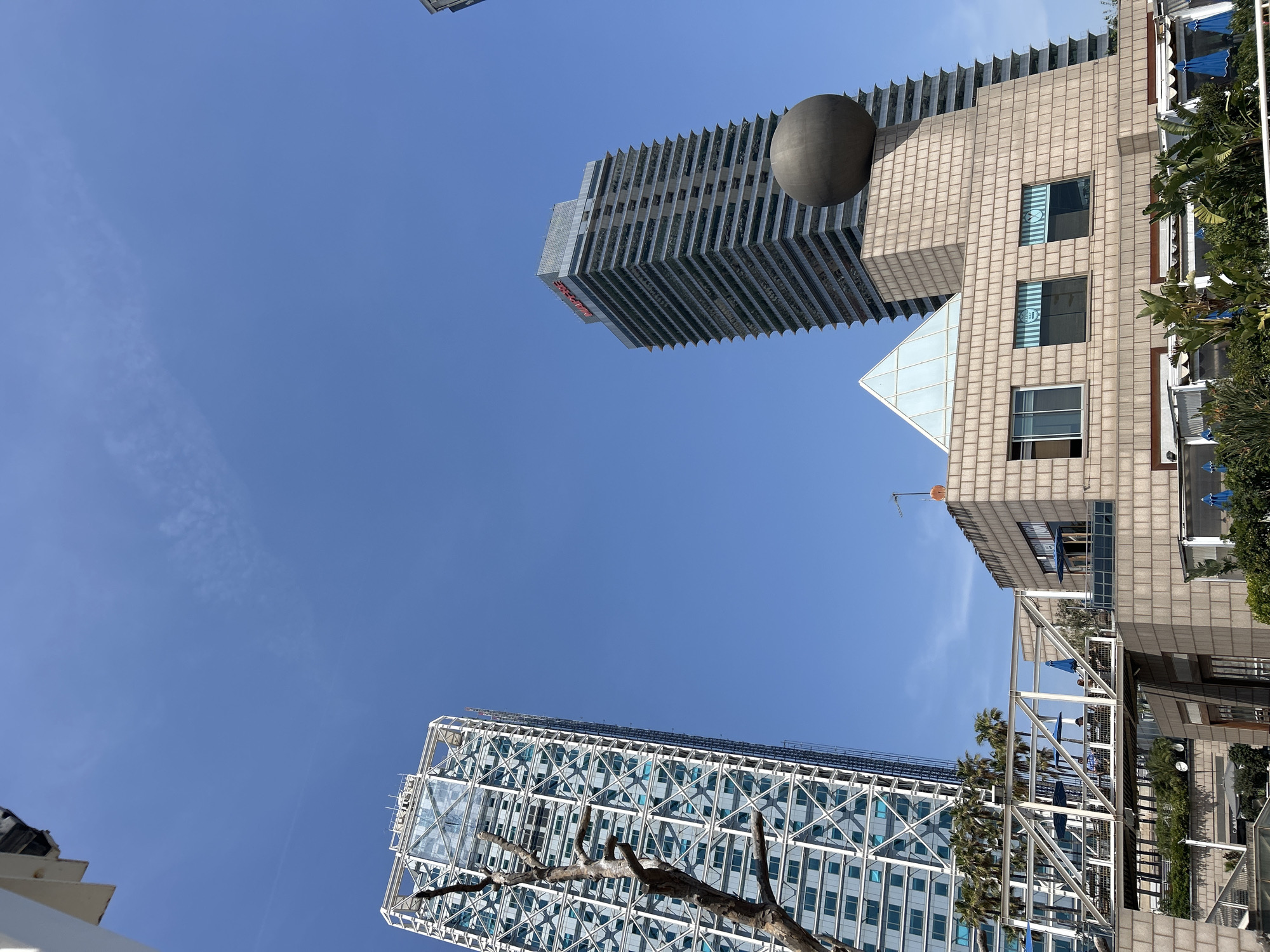 The image shows a low-angle view of buildings in Barcelona, Spain, against a clear blue sky.
The Hotel Arts Barcelona, a modern skyscraper with a distinctive exposed steel structure and a sheer glass facade, is visible in the lower-left corner.
The Hotel Arts Barcelona stands alongside the Torre Mapfre, a skyscraper identifiable by its vertical striations, topped with a large dark-colored sphere.
The third prominent building, situated to the right, is a building of a different architectural style with a light-colored, grid-like facade. This building connects to a series of outdoor spaces and structures, including a terrace or balcony area with tables, chairs, and blue umbrellas.
Trees and some vegetation are visible along the right edge.