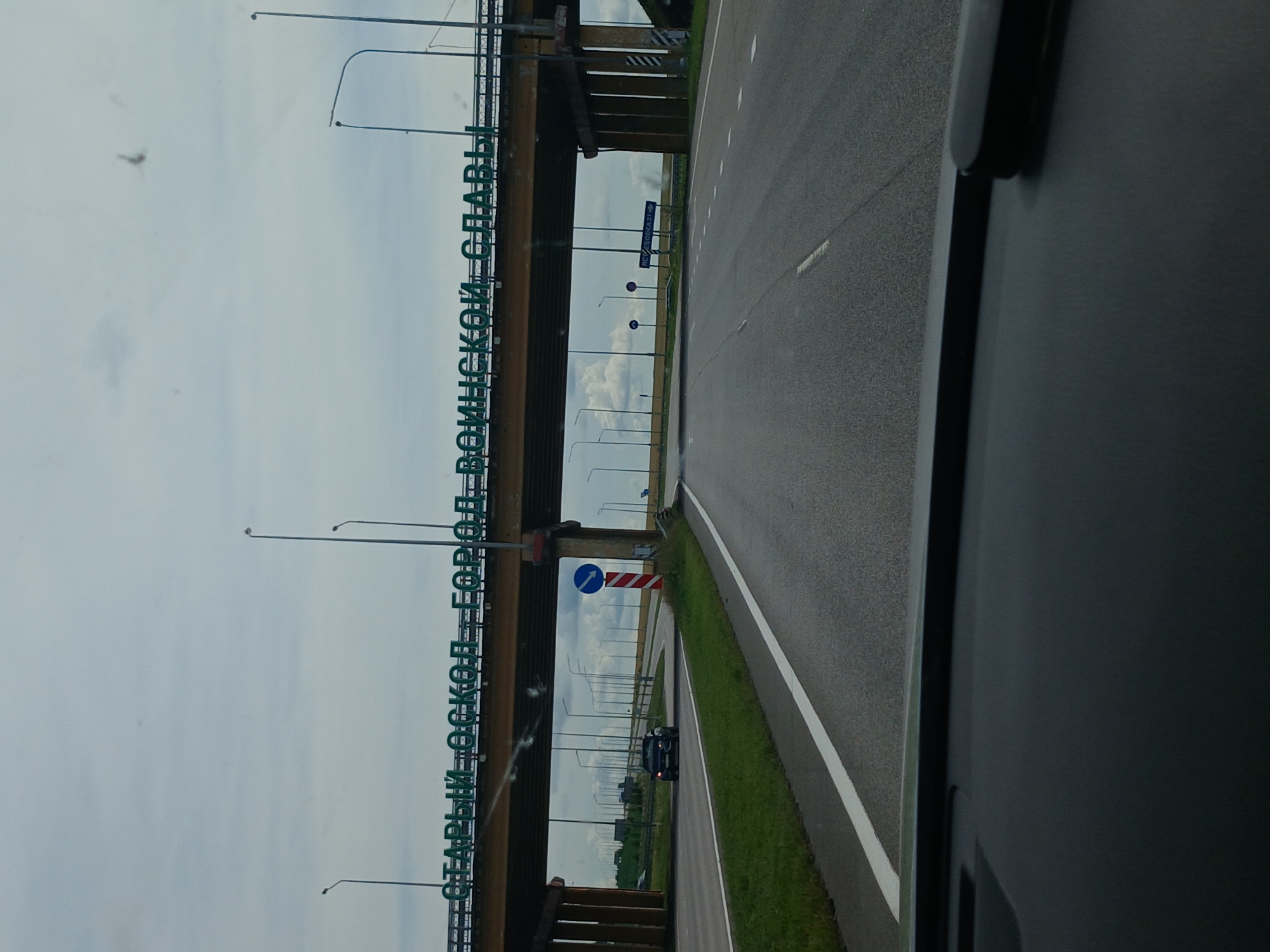The image shows a view from a car approaching a highway overpass with a sign that reads "СТАРЫЙ ОСКОЛ - ГОРОД ВОИНСКОЙ СЛАВЫ" (Stary Oskol - City of Military Glory). The overpass is supported by large concrete pillars and has streetlights along its length. Underneath the overpass, a black car is visible on the road. The surrounding environment includes green grass, fields, and a cloudy sky. There are also road signs, including one with a blue arrow indicating a turn. The perspective suggests the image was taken from the driver's seat of a vehicle.