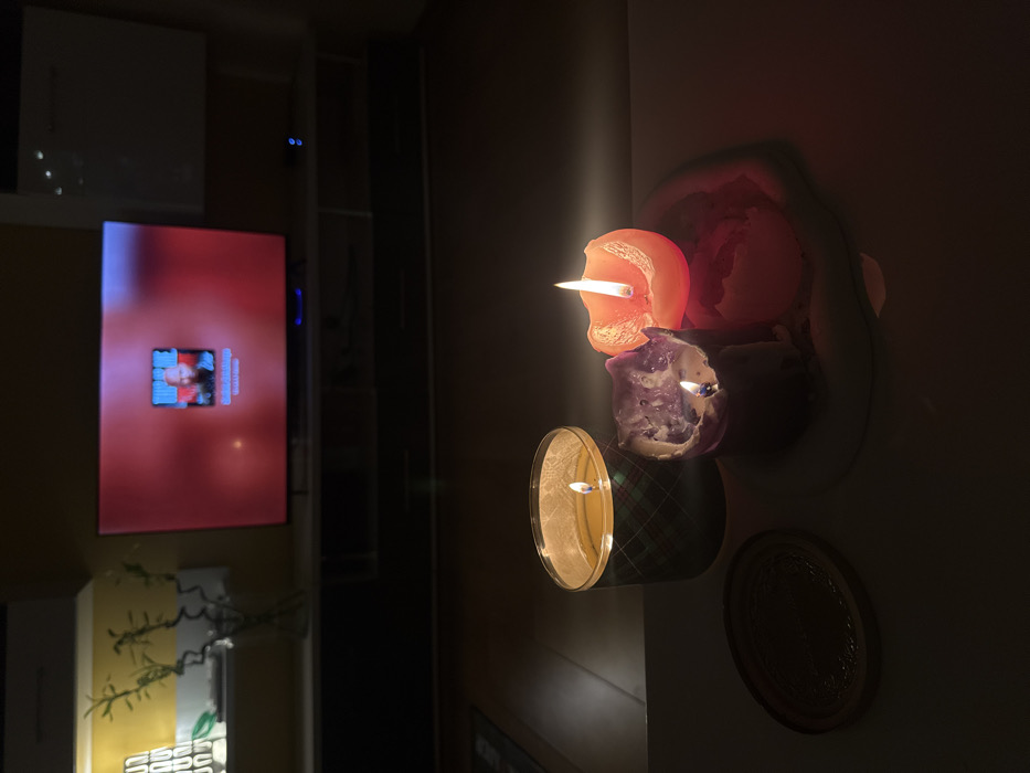 The image shows a dimly lit room with a focus on lit candles arranged on a wall. The top candle is orange, the middle purple, and the bottom one is in a container with a plaid pattern. The flickering flames cast light on the surrounding wall. A television screen with a blurry face is visible on the left, adding a source of ambient light in the background.