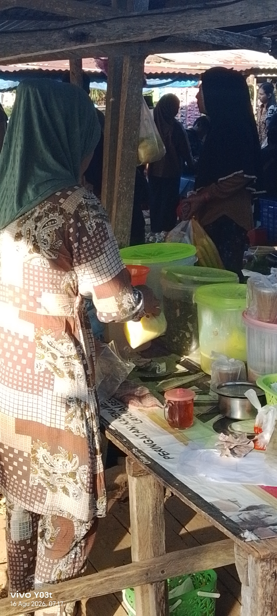 In Palosilae, Indonesia, a scene unfolds at a market stall. A woman wearing a green hijab and a patterned brown and white garment stands in the foreground, her arm extended as she handles a yellow object, possibly a piece of fruit. To her right, several large plastic containers with green lids are visible, some filled with what appears to be dried goods or herbs. A small glass mug filled with a reddish liquid, likely tea or a beverage, sits on the table. A metal bowl and various plastic bags are also present. In the background, other individuals can be seen, some wearing hijabs, suggesting a bustling market environment. The stall is constructed from rough wooden beams and a corrugated metal roof, indicative of a local, open-air market. A printed banner with text, including the Indonesian word "PERINGATAN" (warning), is draped across the front of the stall, partially obscuring items beneath. The lighting suggests it is daytime, with sunlight filtering through the structures.
