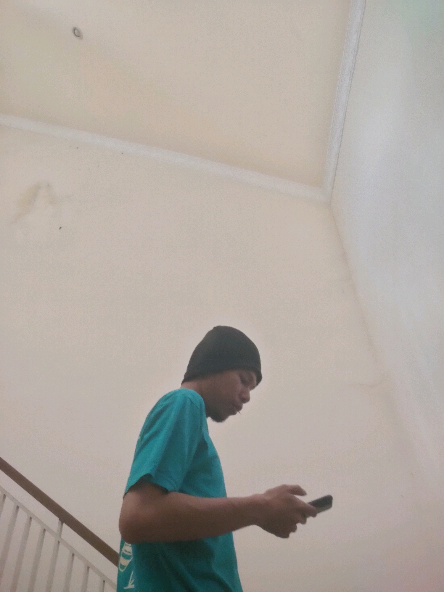 A person wearing a teal t-shirt and a black beanie is standing on a staircase in a building, looking at their phone. The walls of the building are painted a light beige color, and the ceiling is visible at the top of the frame. A wooden handrail runs along the left side of the staircase. The image appears to be taken indoors, possibly in a residential or public building in Sidoarjo, Indonesia.