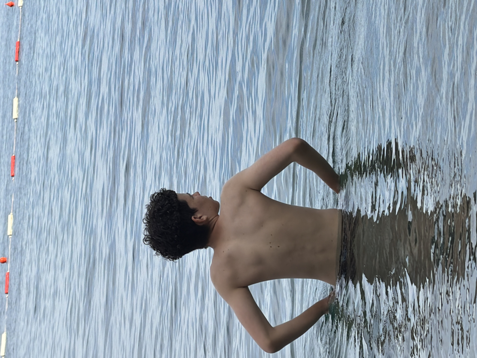 The image depicts a young person, seen from the back and side, standing in shallow, rippling water. Their head is turned to the left, with curly dark hair. They are shirtless, with their arms bent and extended outwards, hands submerged in the water. The lower half of their body is also in the water, wearing patterned swim trunks. The water reflects the sky and surroundings, creating a distorted, watery surface.
To the left of the frame, a rope with alternating red and white buoys is visible, suggesting the edge of a swimming area or a boundary marker. The overall scene appears to be outdoors, likely at a lake, beach, or pool, given the presence of buoys and the body of water. The lighting suggests it is daytime, with soft, diffused light. The ripples on the water indicate a slight breeze or movement. The person's posture and the way they are interacting with the water might suggest a moment of relaxation, playfulness, or simply enjoying the coolness of the water.
The location context of Budva, Montenegro, places this scene in a coastal region known for its beaches and water activities, aligning with the visual cues in the image. No visible text is present in the image.