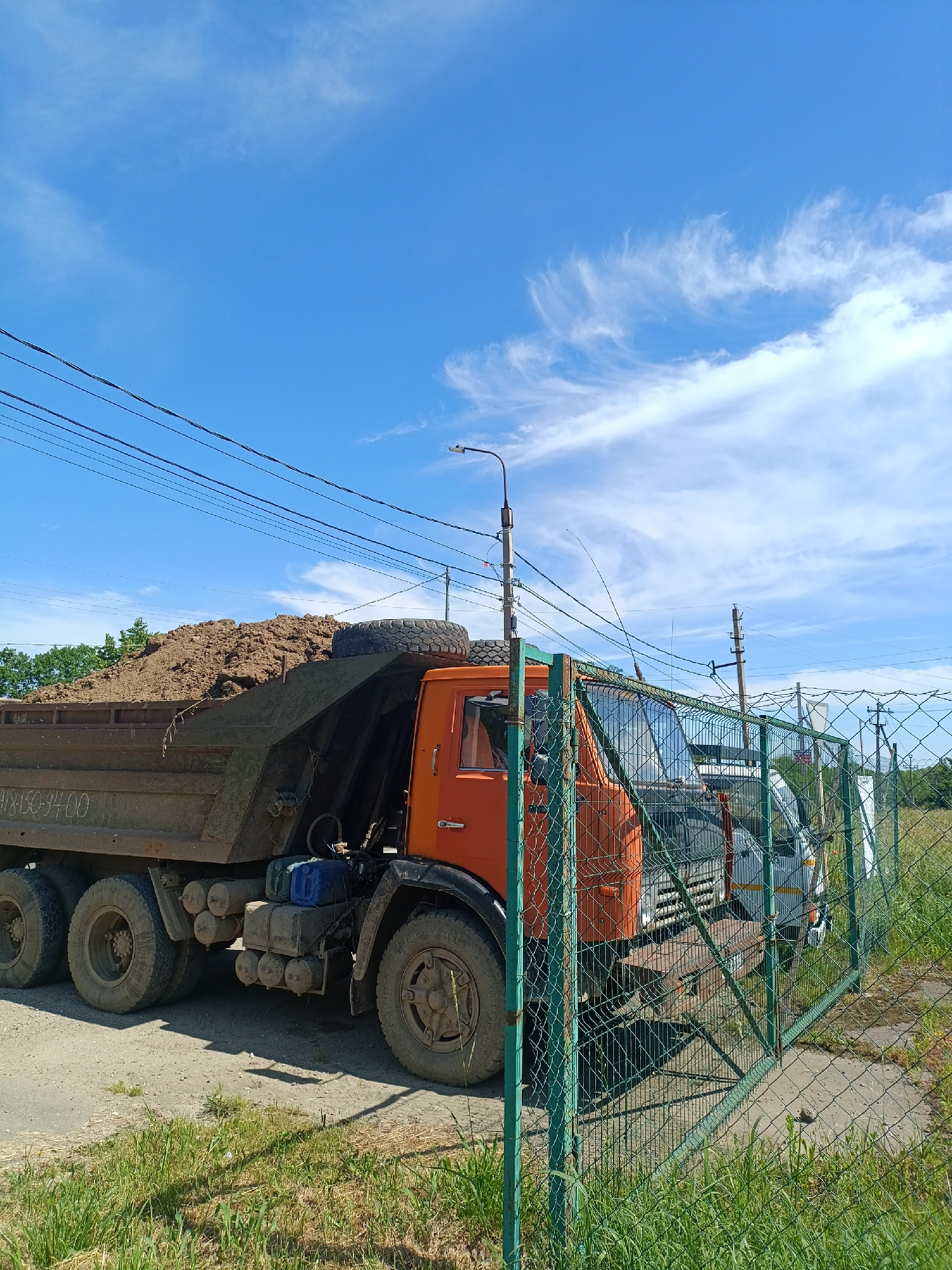 An orange dump truck loaded with soil is parked on a dirt road next to a green chain-link fence. The truck has multiple tires and a visible license plate with the numbers 418150-94. Behind the truck, other vehicles are visible through the fence. The sky is blue with scattered white clouds, and there are power lines overhead. Green grass and trees are present in the background. This scene appears to be in Goryachiy Klyuch, Russia.