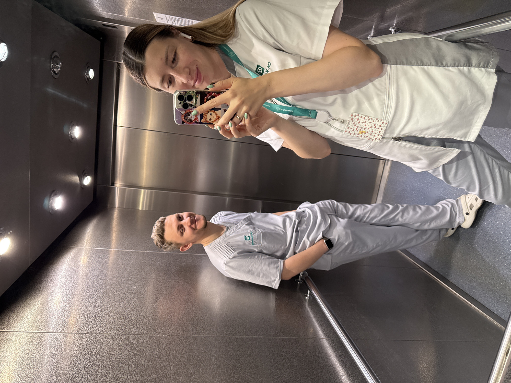 Two individuals, dressed in medical scrubs, are captured in a selfie taken inside an elevator. The image is rotated 90 degrees clockwise. The setting is an elevator with metallic, reflective walls and a dark, textured floor. Several recessed lights illuminate the ceiling.
In the foreground (top of the rotated image), a female with long brown hair, wearing a white scrub top with light grey accents and light grey pants, is taking the selfie. She holds an iPhone with a colorful, illustrated phone case (featuring cartoon-like characters) in her right hand. She has mint green nail polish and a wedding band on her left ring finger. White earbuds are visible in her ears. Around her neck, she wears a teal lanyard with the text "CW-MD" and a logo that appears to be a medical cross or stylized symbol. An ID card holder with a pattern of small red and orange shapes is clipped to her top. She is looking at her phone screen, smiling faintly.
Reflected in the elevator wall to her left (right in the rotated image), a male with short blonde hair stands, also smiling faintly. He is wearing light grey scrubs, with a green logo (likely matching the "CW-MD" logo) visible on his left chest pocket. He has a dark smartwatch on his left wrist and wears white Crocs on his feet. A metal handrail is visible on the wall behind him.
Both individuals appear to be healthcare professionals, likely at a medical facility in Moscow, given the provided context and their attire. The activity is a casual selfie moment within their workplace environment.