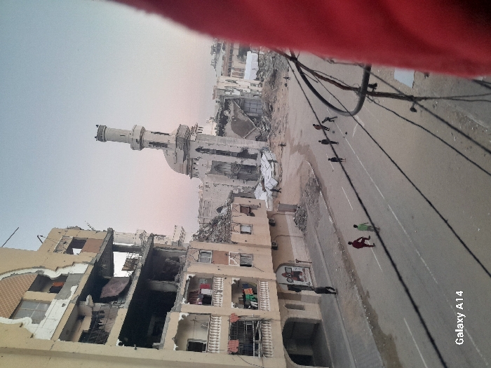 The scene depicts a war-torn urban landscape. Buildings are severely damaged, some reduced to rubble. A mosque with a prominent minaret stands amidst the destruction. A road cuts through the scene, where several individuals are walking. Overhead power lines crisscross the sky. The overall impression is one of devastation and hardship. The photograph has a red tinge at the top edge of the image. The bottom right corner has the text Galaxy A14.