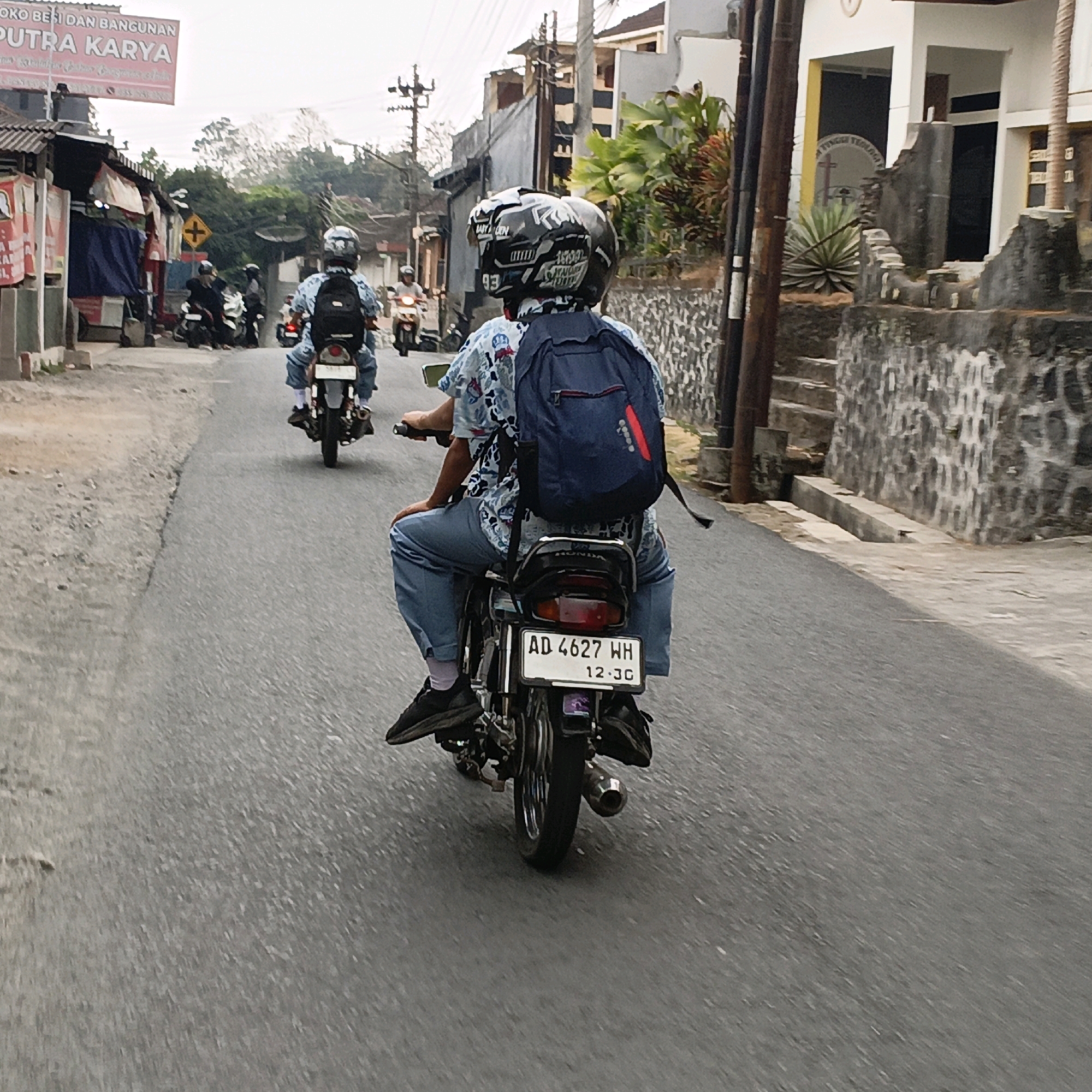 In Salatiga, Indonesia, a group of individuals ride motorcycles down a street lined with buildings and trees. The primary focus is on the motorcyclist in the foreground, wearing a blue patterned shirt and jeans, with a blue backpack. They are riding a black motorcycle with a license plate that reads "AD 4627 WH 12-26". Behind them, other motorcyclists are also visible, some with backpacks and helmets. The street is paved, and there is a slight incline. The buildings on either side of the street vary in architectural style and condition, with some appearing to be shops or houses. The sky is overcast, suggesting a cloudy day. The scene captures a typical street view in Salatiga, showcasing everyday transportation and urban landscape.