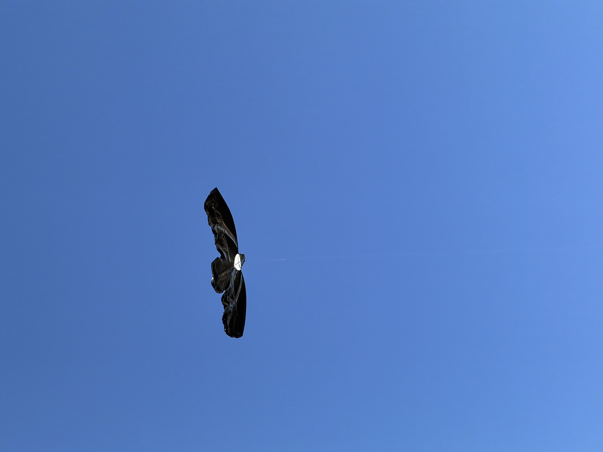 A black kite is in flight against a clear blue sky. The kite is shaped like a manta ray with its wings spread wide. A thin string connects the kite to an unseen point below. The kite appears to be homemade, possibly constructed from plastic. The sky is a uniform blue, suggesting a sunny day. There are no other objects or people visible in the frame. The image was taken in Mengwi, Indonesia.
