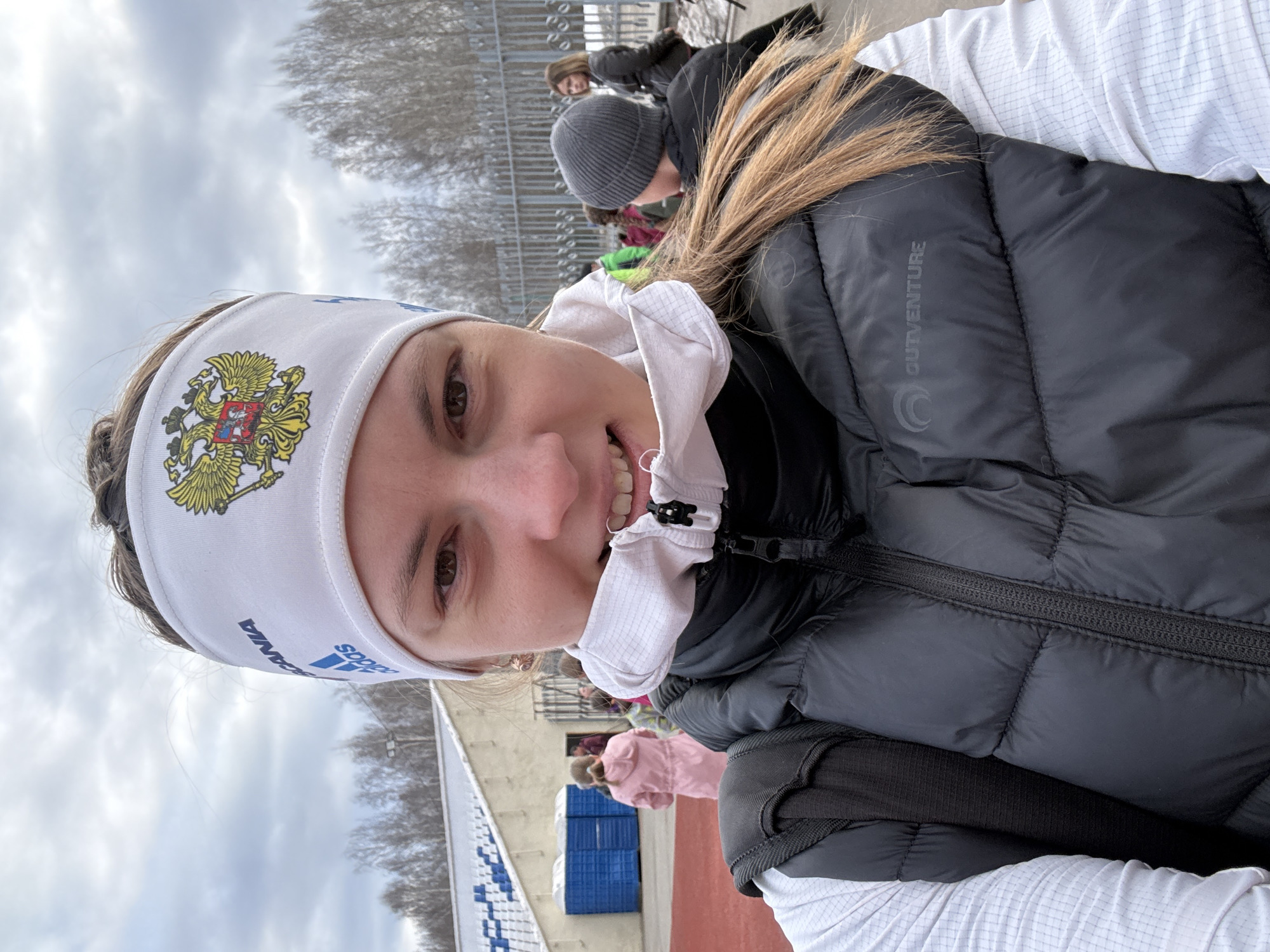 A woman is taking a selfie outdoors in what appears to be a sports or recreational area. She is wearing a white headband with the Russian coat of arms. The headband also has "RUSSIA" and "ADIDAS" written on it. The woman is also wearing a black puffer jacket over a white top.
In the background, there are other people, including one wearing a gray beanie. There's also a fence, trees, and what looks like a stadium with blue seats. The sky is overcast.
Given the headband and stadium in the background, along with the reported location of Chaikovsky, Russia, it's possible this photo was taken at the Snezhinka ski complex or another similar sporting venue in the city.