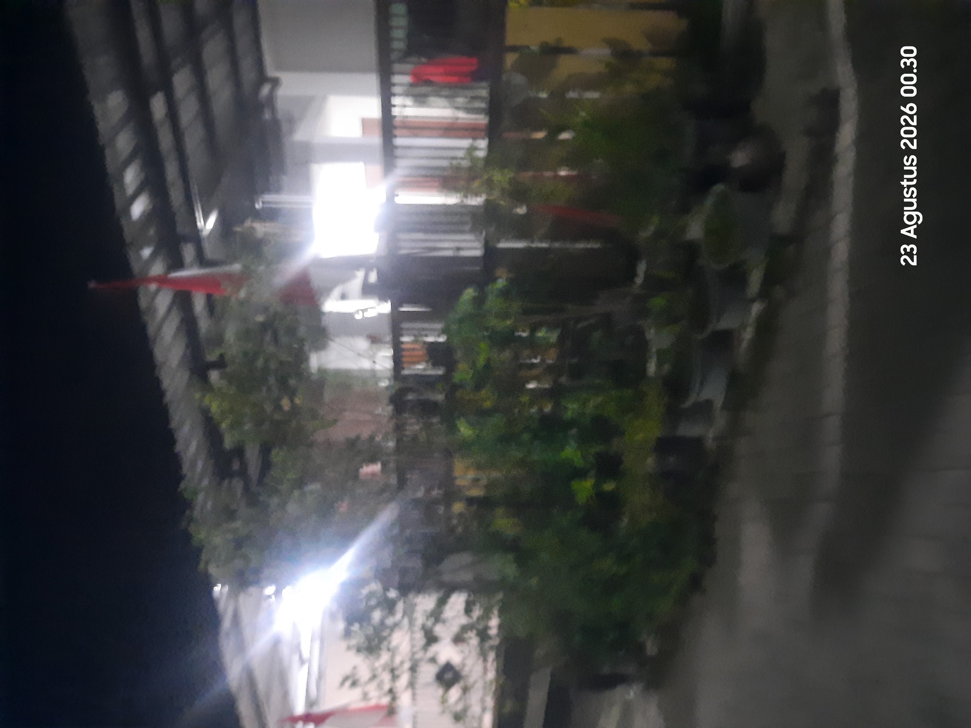 The image shows an outdoor scene at night, with a building and a fence in the background, and several potted plants in the foreground. A red and white flag, possibly the Indonesian flag, is visible on the left side of the building. The overall atmosphere is dim, with bright lights from the building illuminating parts of the scene. A timestamp in the bottom right corner reads "23 Agustus 2026 00.30". The location is identified as Sukokerto, Indonesia.