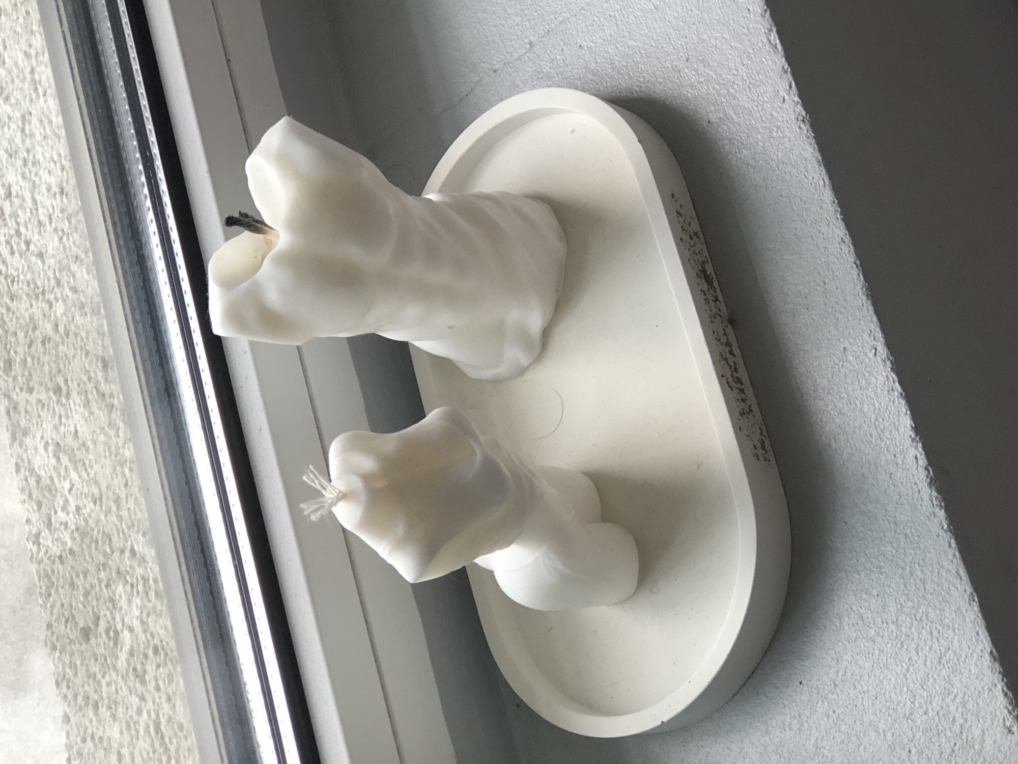 Two white, torso-shaped candles, one male and one female, sit on a cream-colored oval tray. The male torso candle is on the right and appears to have been lit, as its wick is charred. The female torso candle is on the left, with an unlit, frayed wick. The tray is placed on a grey, textured surface next to a window with a dark frame. The background outside the window is blurred and appears to be a light-colored, textured wall. The scene is lit by natural light coming from the window. The overall setting suggests an indoor display, possibly on a windowsill or shelf. No specific landmarks or context are visible to indicate the city of Saint Petersburg, Russia.