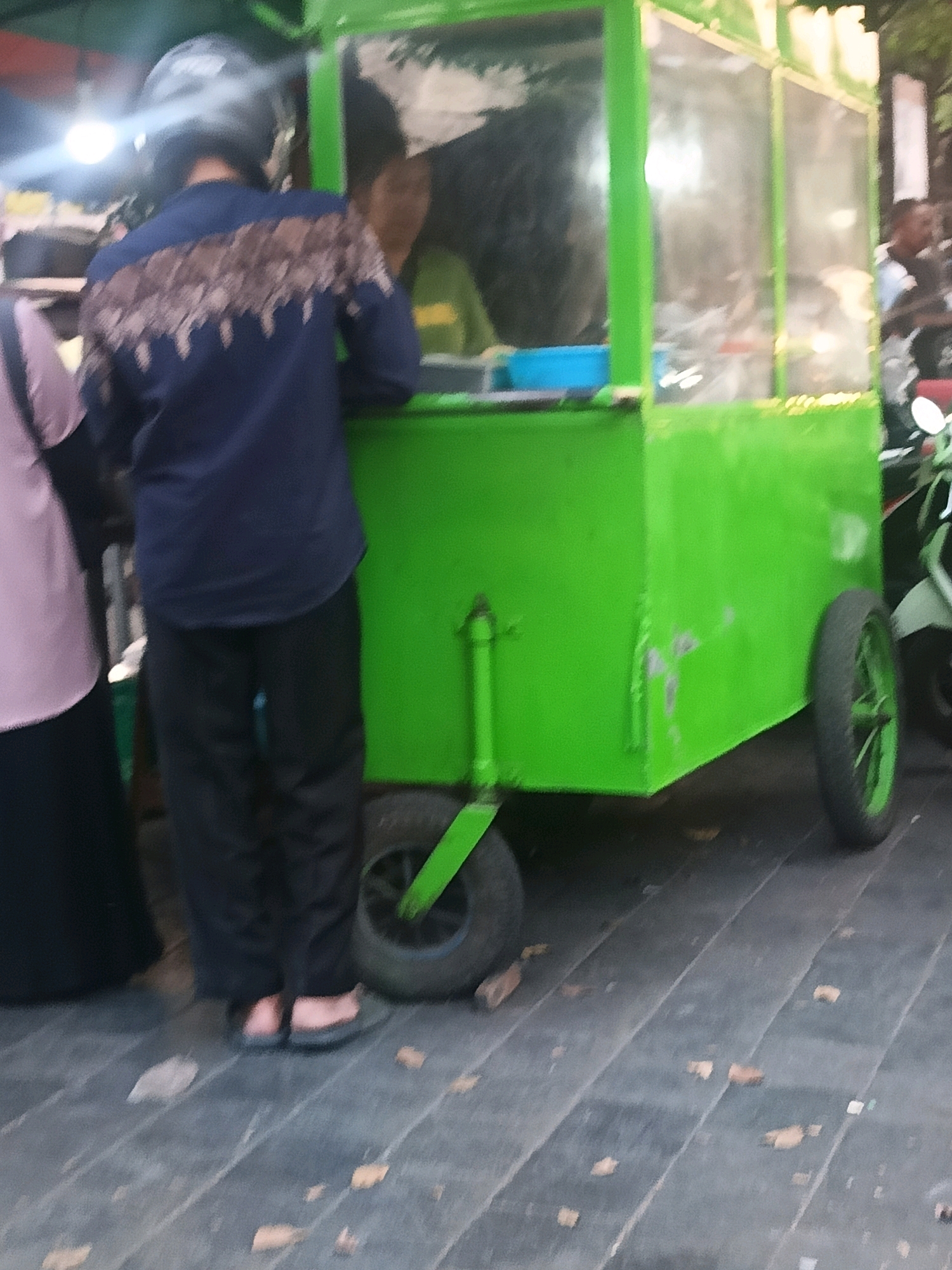 A blurry, medium-shot image shows a person wearing a dark blue shirt with a patterned design and black pants, standing next to a bright green food cart. The person is wearing a dark helmet and dark flip-flops. Another person, wearing a light purple shirt and black pants, is partially visible on the left side of the frame. The food cart has glass windows and wheels. The ground is paved with dark wood-like planks, and a few fallen leaves are scattered on the ground. The scene appears to be outdoors, possibly in a public area in the city of Yogyakarta, Indonesia, given the style of the food cart and the commonality of street food vendors in the region.