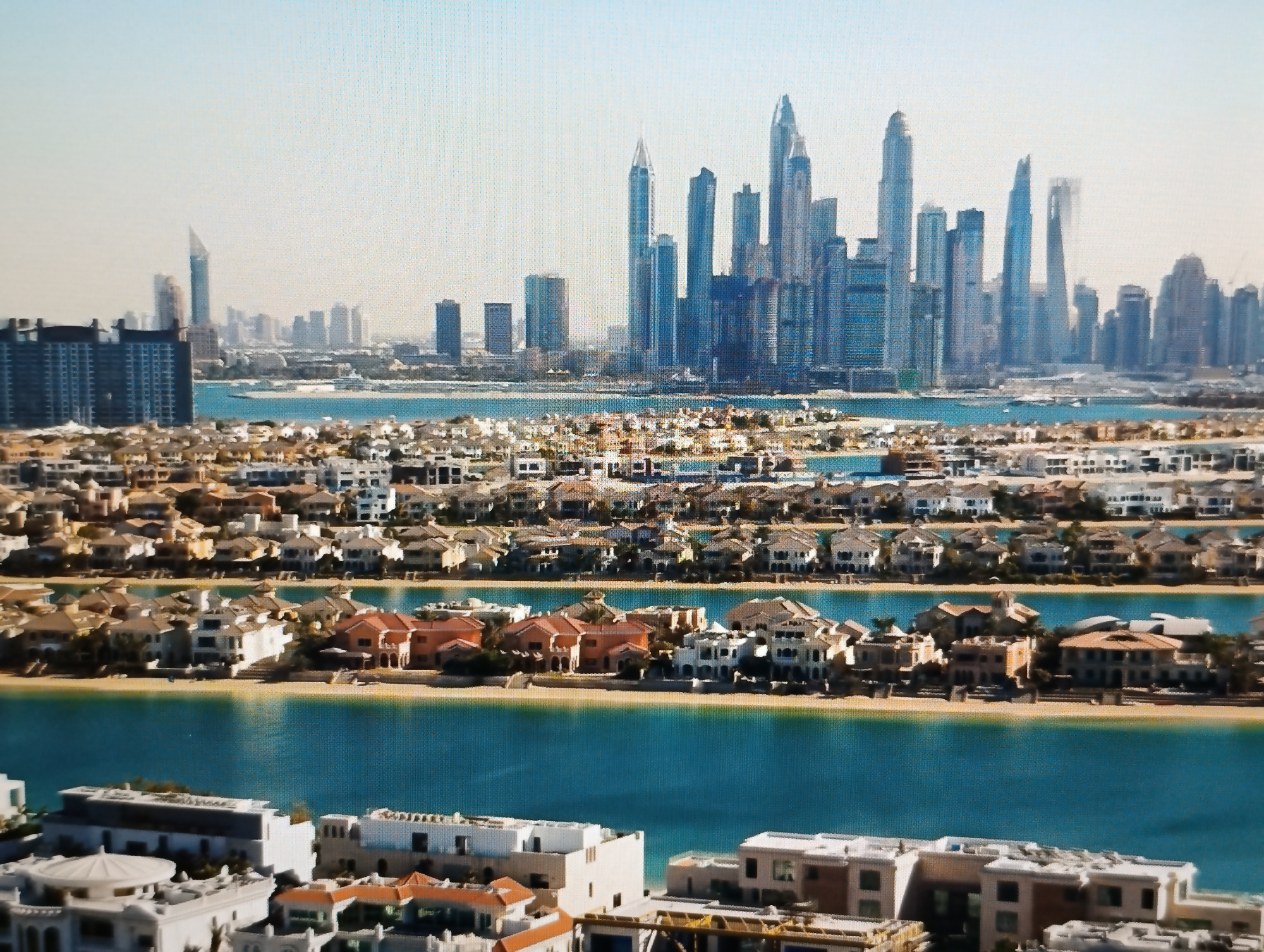 The image presents a wide-angle aerial view of a coastal cityscape with a distinct architectural style. In the foreground, a cluster of multi-story buildings with flat roofs and white facades lines the waterfront. Beyond this, a series of parallel rows of modern villas, characterized by terracotta roofs and sandy-colored walls, are situated along a bright turquoise body of water. Further in the distance, a dense collection of towering skyscrapers dominates the horizon, indicating a major urban center. The sky above is a pale, hazy blue. The scene appears to be a resort or residential area with a modern, affluent aesthetic. The query about the location of "Dubăsari town council, Moldova" is irrelevant to the visual content of the image, as the architecture and cityscape depicted are clearly not characteristic of Moldova.