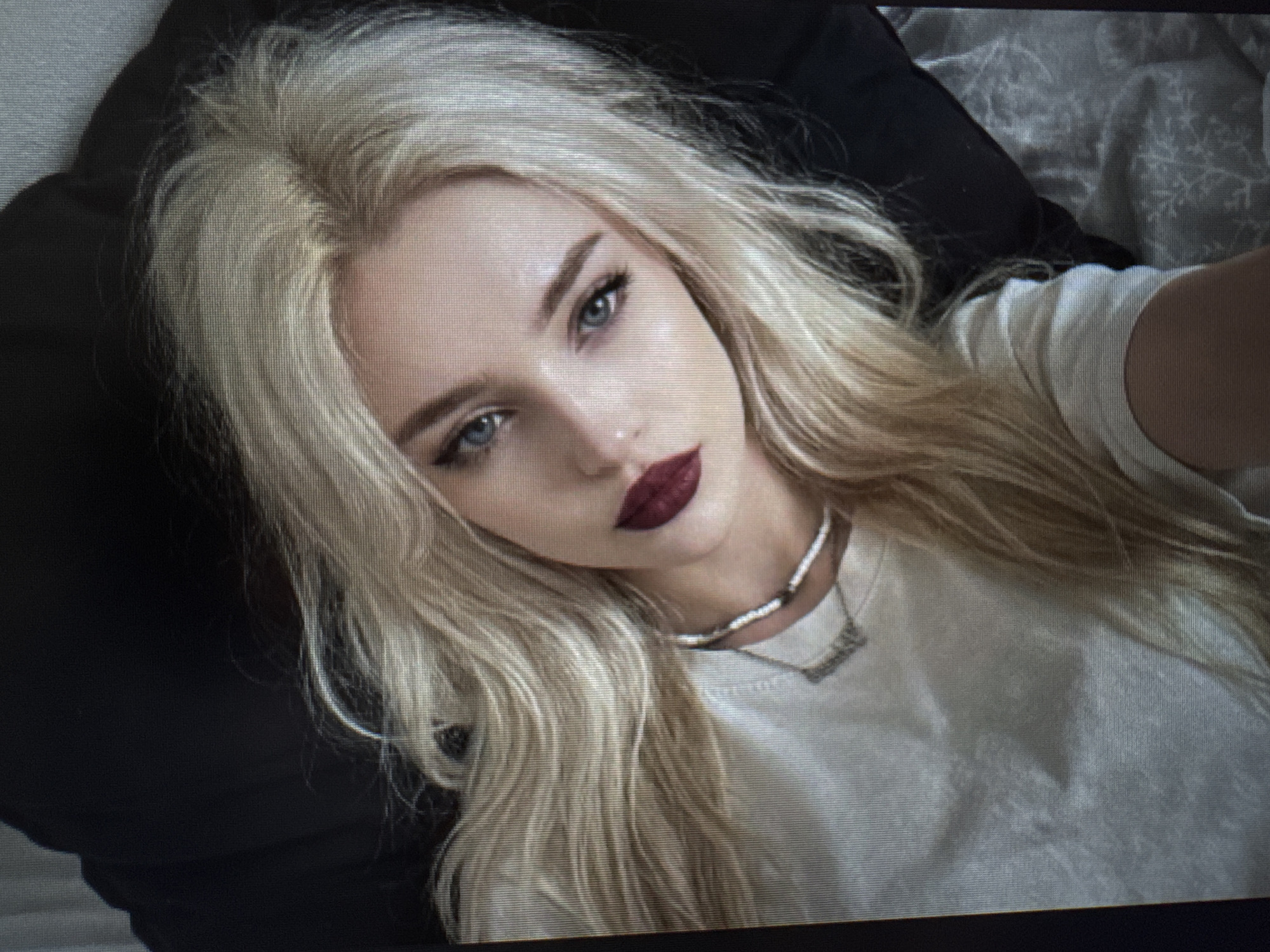 A young woman with platinum blonde hair and striking blue eyes is depicted in a close-up, eye-level shot, appearing to be a selfie. She has defined eyebrows, winged eyeliner, and dark, matte burgundy lipstick. Her long, wavy blonde hair shows visible darker roots near the scalp.
She is wearing a simple white crew-neck t-shirt. Around her neck are two necklaces: a beaded choker made of small white or light-colored beads, and a longer, delicate silver-toned chain with a small, unidentifiable pendant. The pendant appears to have text, but it is too small and pixelated to be legible.
The woman is positioned against a dark black pillow or fabric on the left side of the frame, and a grey and white patterned fabric, possibly a blanket or sheet with a snowflake-like design, on the right. Her posture suggests she is lying down, looking directly at the camera. The overall lighting is soft and even, typical of indoor conditions.
A notable detail of the image itself is the visible horizontal lines and pixelation, particularly evident in the cropped sections, indicating it is likely a photograph of a digital screen rather than a direct digital image.