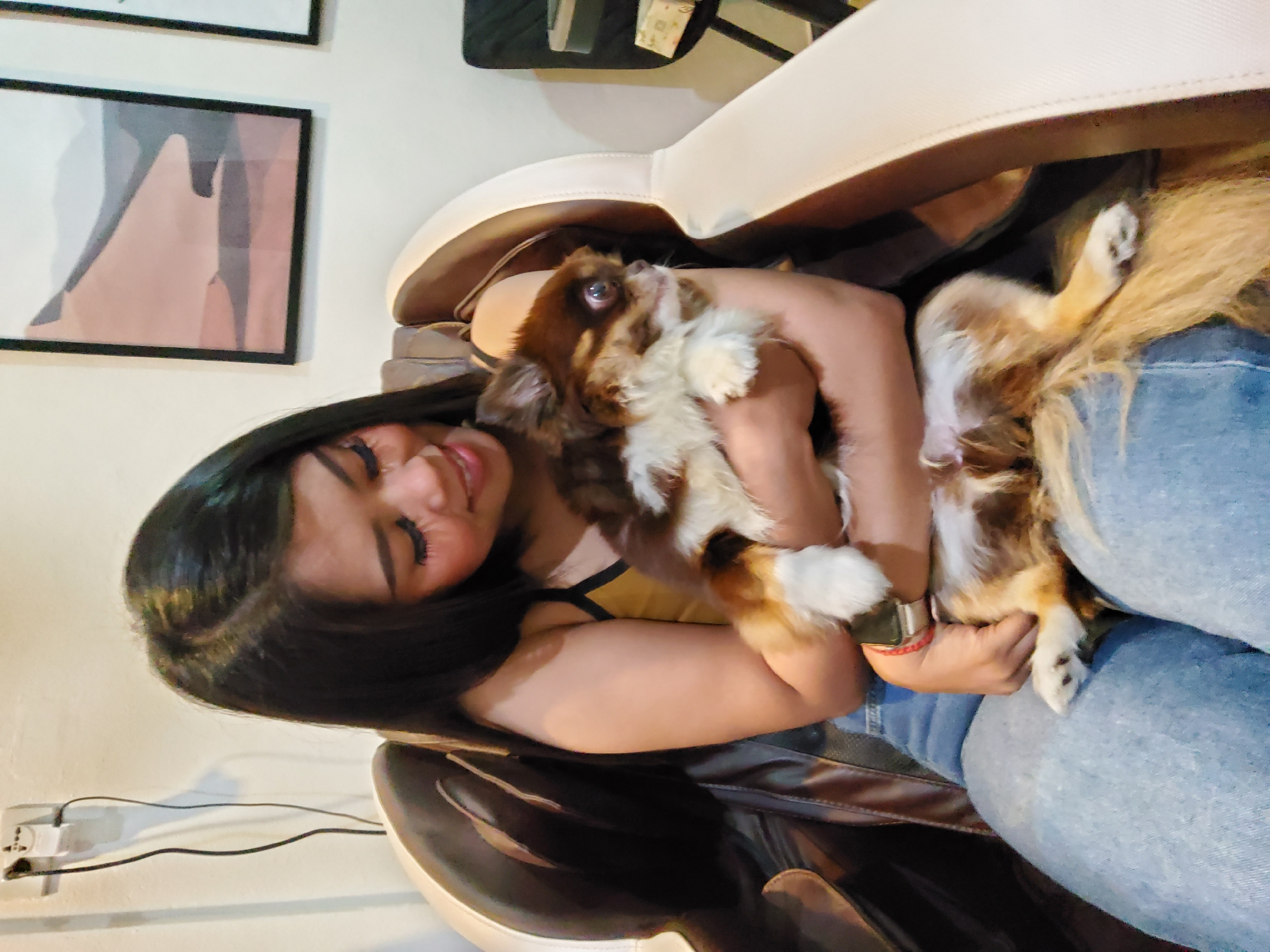In the city of Trang, Thailand, a young woman with long, dark hair and dramatic eye makeup sits in a brown and cream massage chair. She is holding a small, long-haired chihuahua. The dog is mostly brown with white and tan markings. The woman is wearing a yellow tank top and blue jeans, and a red bracelet is visible on her wrist. The background features a white wall with an electrical outlet and cord, and two framed abstract art pieces depicting layered, muted colors. The overall scene appears to be indoors, likely in a home or a spa setting.