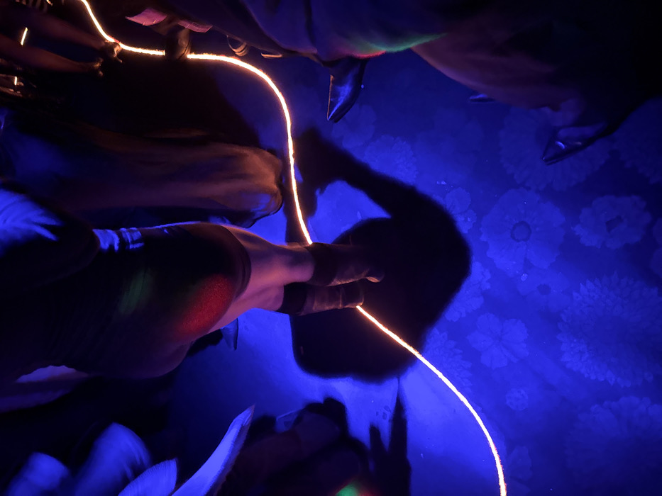The image depicts a crowded interior scene within the Le Balajo nightclub in Paris, France. The environment is illuminated predominantly by deep blue ultraviolet (UV) light, causing a distinct pattern of large white floral designs to luminesce on the dark blue floor. Multiple individuals are present, observed from an overhead perspective, with only their lower torsos, legs, and feet visible. A central figure, oriented with their back partially towards the viewer, wears a dark garment and dark boots/shoes, their substantial shadow cast onto the floor. A red light source reflects distinctly on this individual's right upper thigh. An orange, glowing, curvilinear wire snakes across the floor, passing between the feet of the various attendees. Partial views of other legs and footwear, including pointed-toe shoes, are visible to the left and right, with occasional green light reflections appearing on other subjects. The scene conveys a dynamic, crowded atmosphere, characteristic of a dance floor or social event.