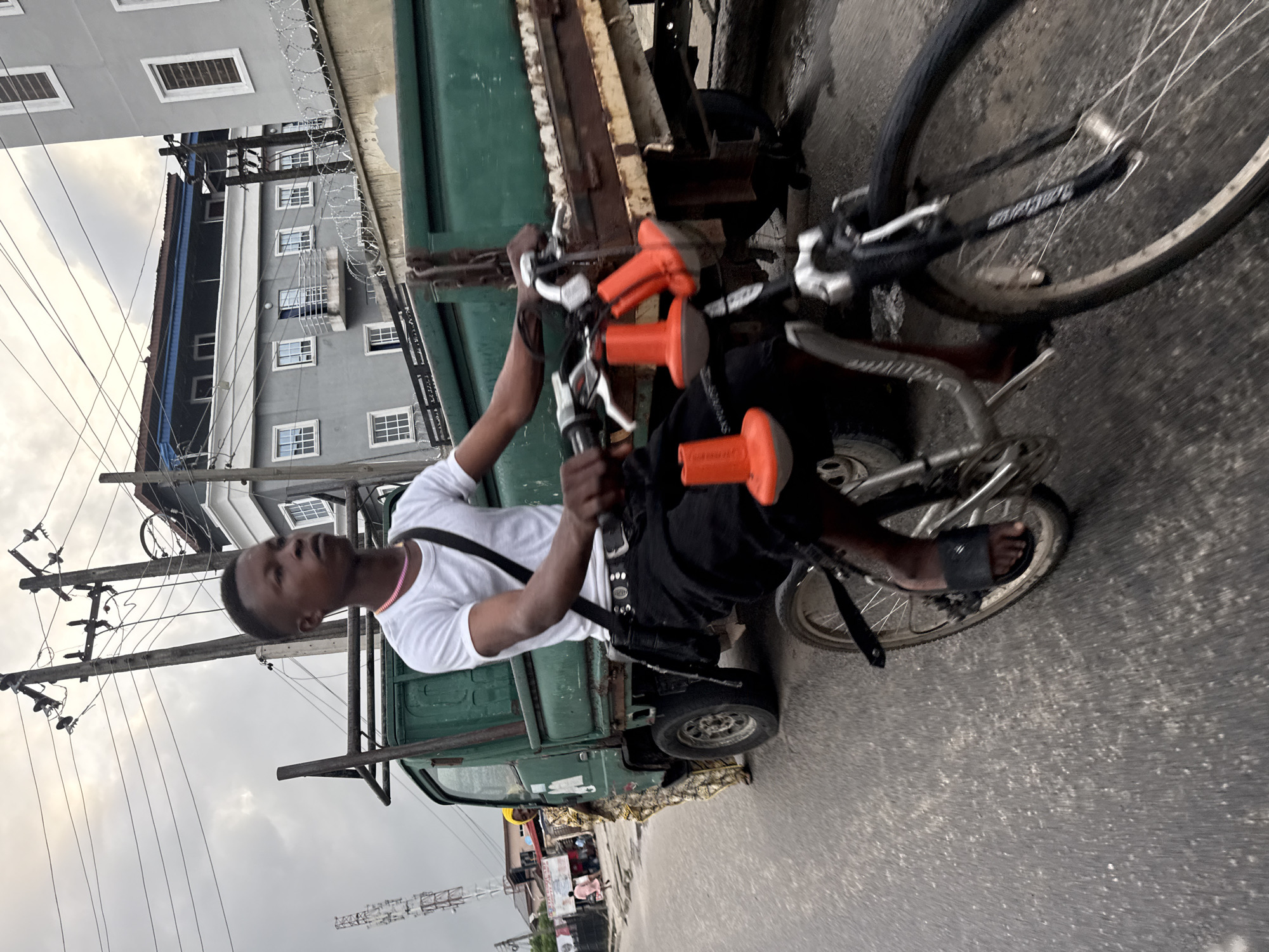 A young man rides a bicycle on a paved road in Ado, Nigeria. He wears a white t-shirt, black pants, and black sandals. The bicycle has two orange cones attached to the handlebars. Behind him is a green truck. In the background are buildings and utility poles with electrical wires. The sky is overcast.