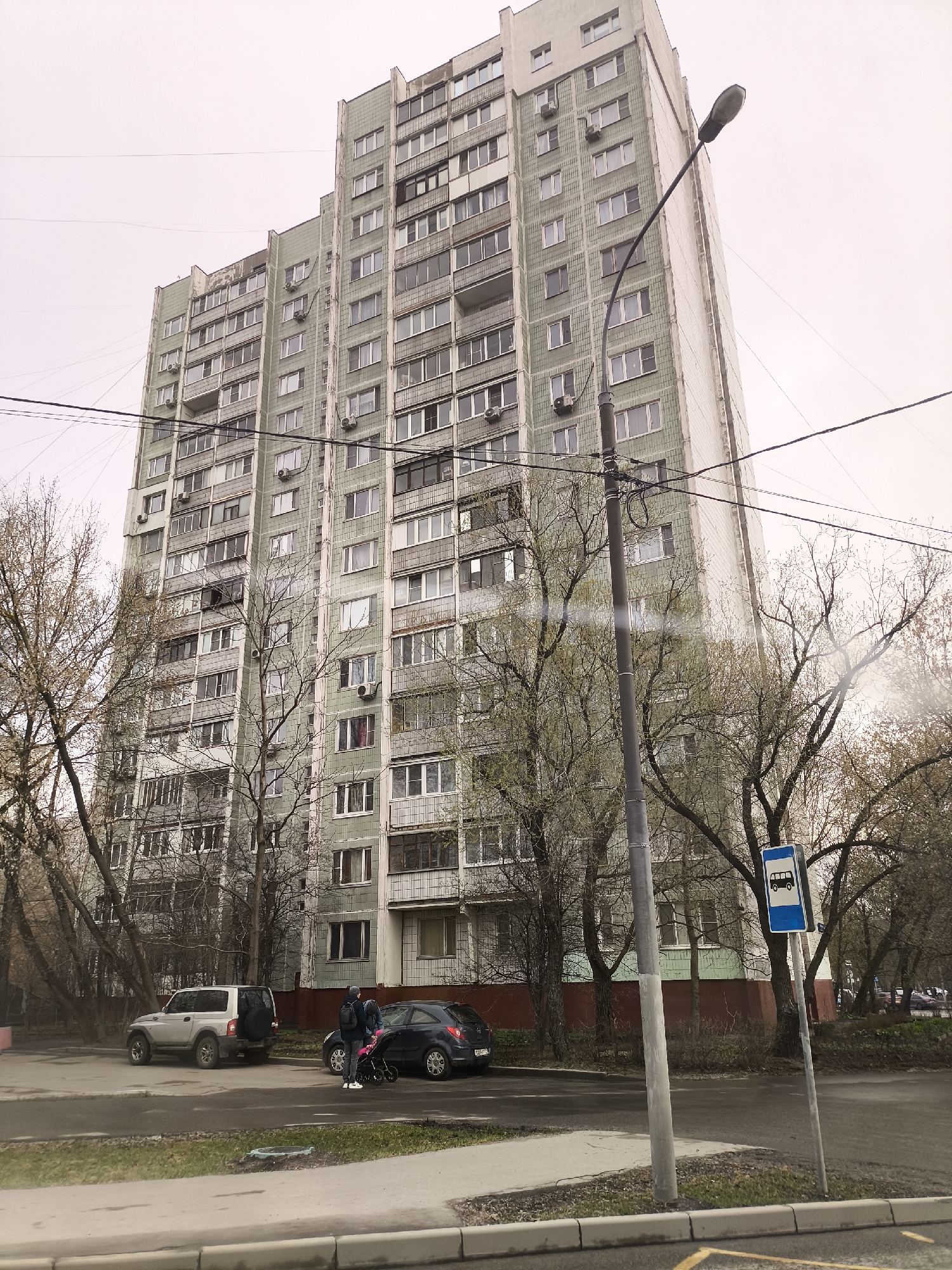 The image depicts a typical urban scene in Moscow, Russia, showcasing a tall, Soviet-era apartment building. The building is a high-rise structure, likely constructed of prefabricated concrete panels, with a predominantly green and gray facade. Rows of windows line the building's exterior, and many have air conditioning units mounted below them.
In the foreground, a street is visible, with parked cars including a silver SUV and a dark-colored hatchback. A couple stands on the sidewalk next to the hatchback, and they are accompanied by a baby stroller. A bus stop sign is prominently positioned on the right side of the frame, indicating the presence of public transportation. Bare trees are scattered throughout the scene, suggesting it is either late autumn or early spring. Overhead power lines crisscross the sky, adding to the urban feel of the setting. The overcast sky contributes to the muted color palette of the image.