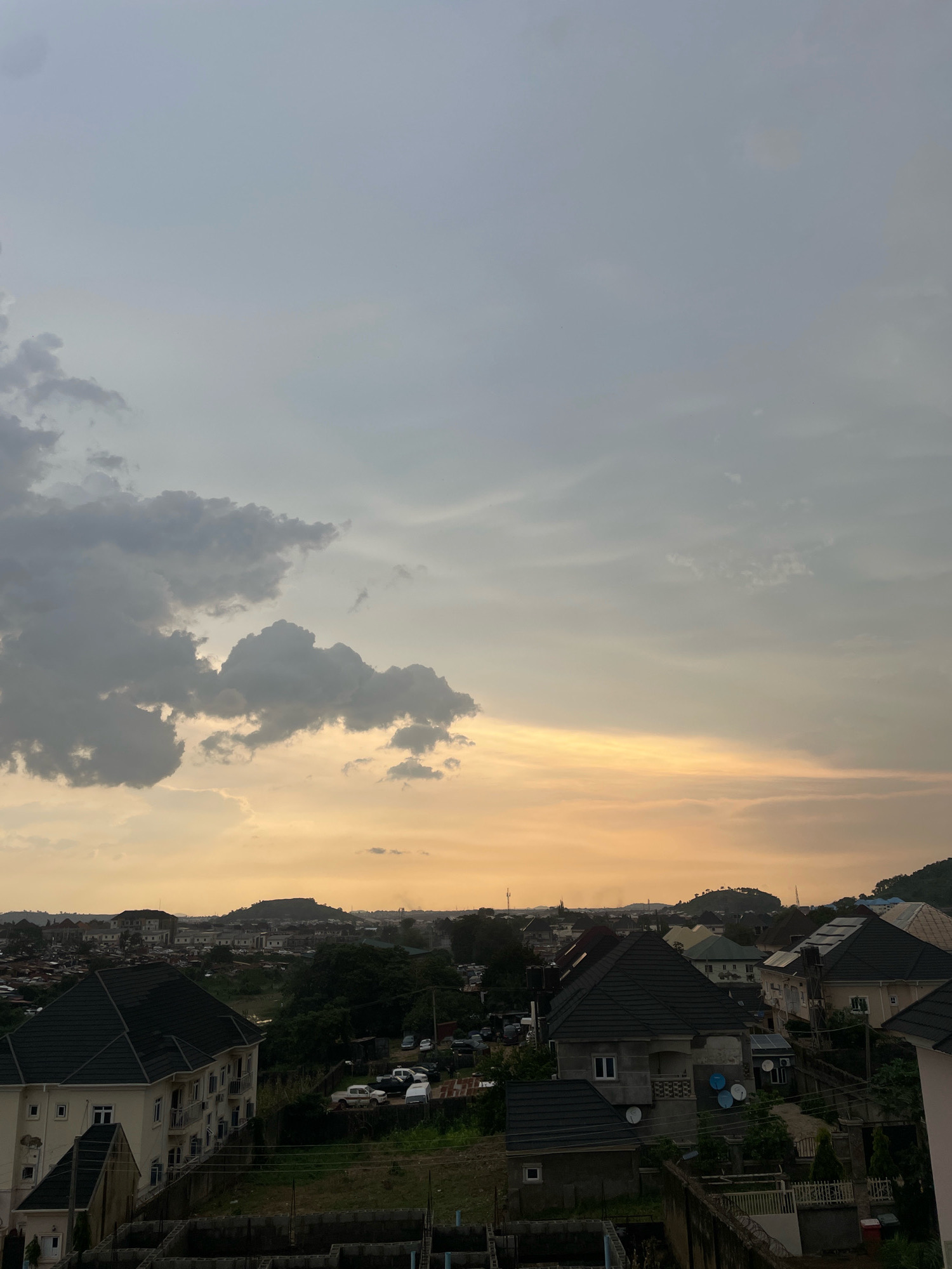 The image depicts an aerial view of a residential area in Abuja, Nigeria, at dusk. The sky is filled with dramatic clouds, with a warm, orange glow visible near the horizon, suggesting the sun is setting. In the foreground, several buildings with dark-tiled roofs are visible, along with some construction sites. A larger, cream-colored building with multiple stories stands prominently on the left, showcasing a modern architectural style. To the right, smaller houses with various roof designs are clustered together, with some featuring satellite dishes. The middle ground shows a densely packed urban landscape with trees and more buildings, interspersed with parked vehicles. In the background, rolling hills and a distant mountain range are silhouetted against the twilight sky, indicating the hilly topography of Abuja. The overall scene conveys a sense of urban development and the natural beauty of the surrounding landscape.