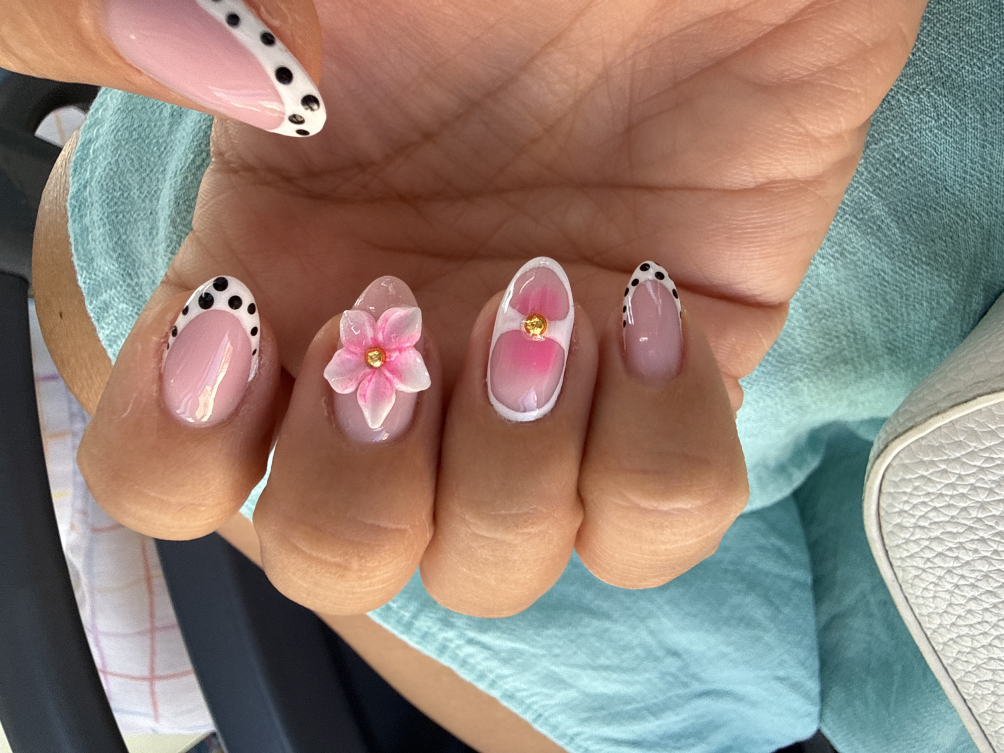 The image shows a close-up of a person's hand with manicured fingernails. The nails are decorated with a French manicure style, featuring a pink base with white tips. Some nails have black polka dots along the white tips. Two nails feature a 3D pink flower embellishment with a gold stud in the center. The hand is resting on a teal-colored textured fabric, possibly a towel or clothing. In the background, a dark surface and a white and pink checkered pattern are visible. No specific location in Varna, Bulgaria is identifiable from the image.