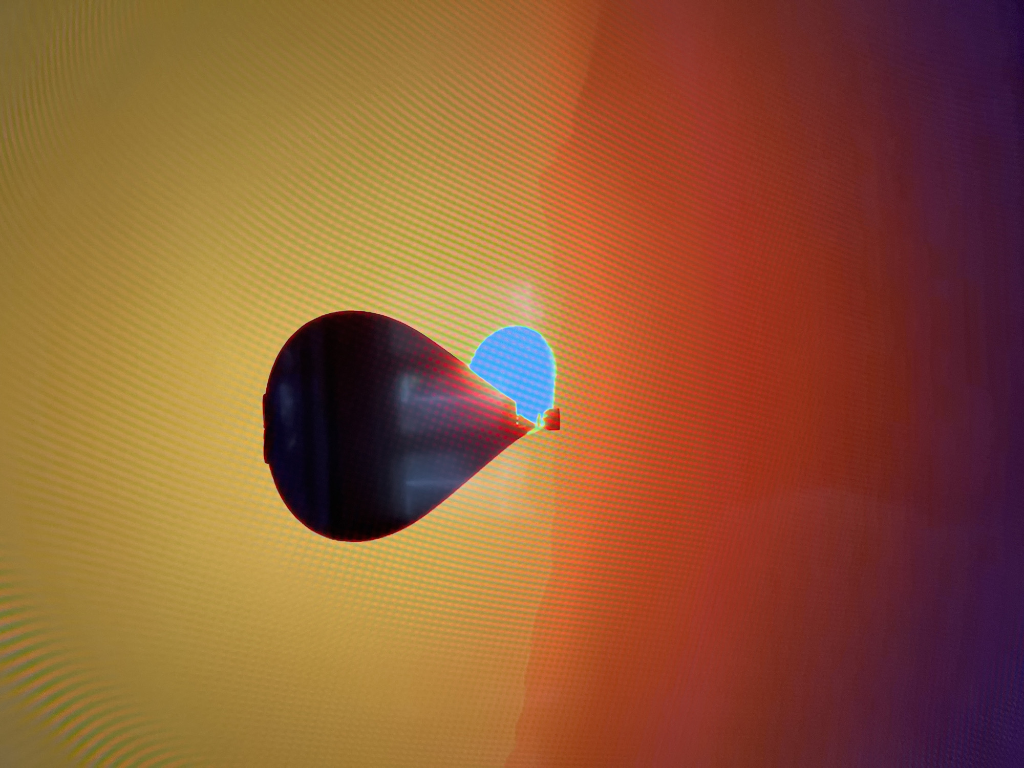A hot air balloon is depicted against a sunset or sunrise sky. The sky transitions from yellow at the top to orange and then purple at the bottom, with a textured appearance possibly due to a digital display. A small, blue object, possibly a cloud or another balloon, is visible near the lower portion of the main balloon. The hot air balloon is dark at the top and shows red and blue hues towards its bottom. The scene is abstract and does not depict any specific identifiable landmarks or details of Kharkiv, Ukraine.