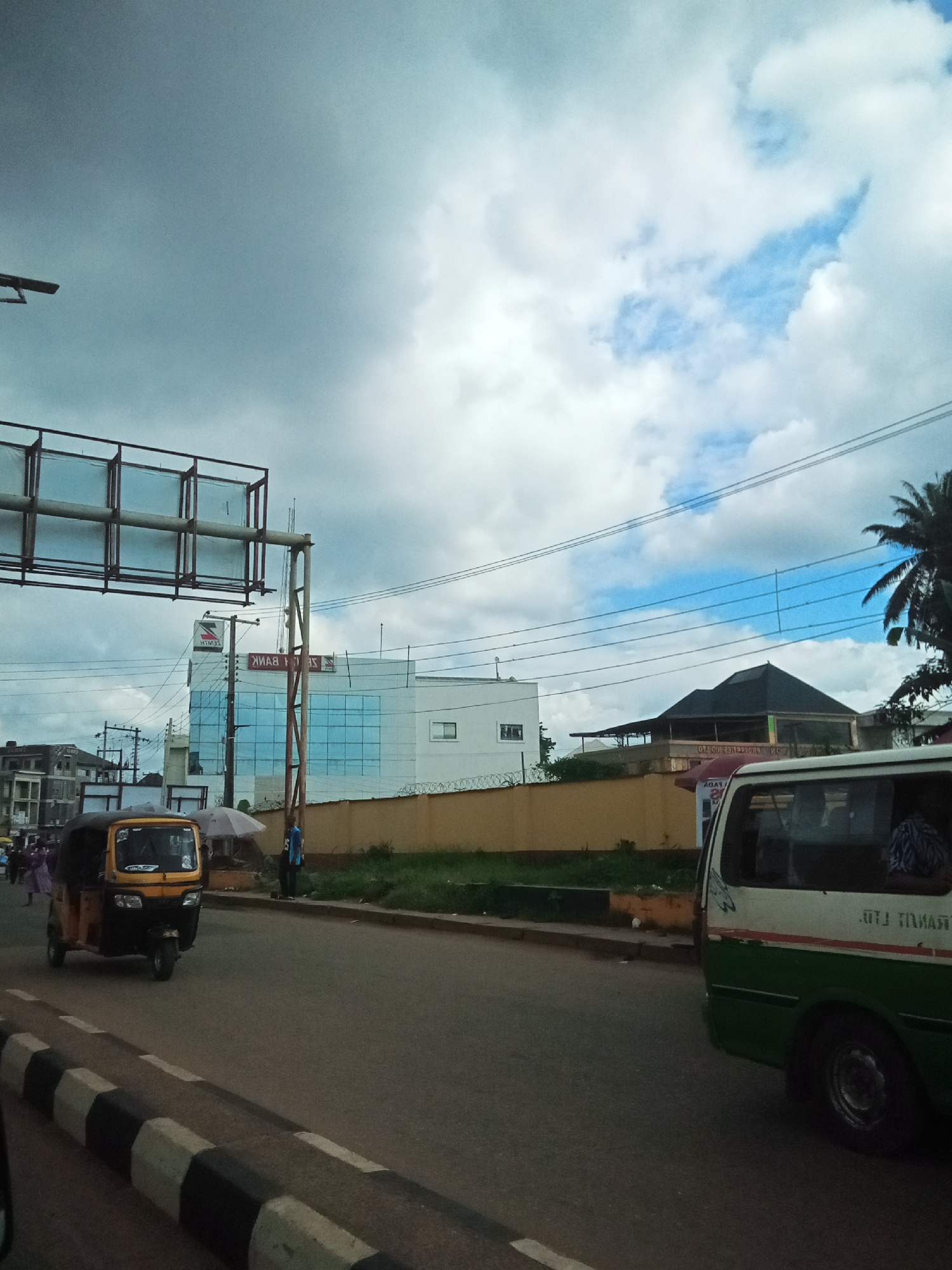 In Nibo, Nigeria, this image captures a street scene with a yellow auto-rickshaw in motion on the left, its driver visible. A white and green bus is parked or moving slowly on the right, showing passengers inside. In the background, a modern building with blue-tinted windows, displaying a "ZENITH BANK" logo, stands prominently. Other structures and trees are visible, and power lines crisscross the sky, which is partially overcast with dramatic clouds. A guardrail or fence borders a yellow wall along the street. The road surface is asphalt, marked with a black and white striped speed bump in the foreground.