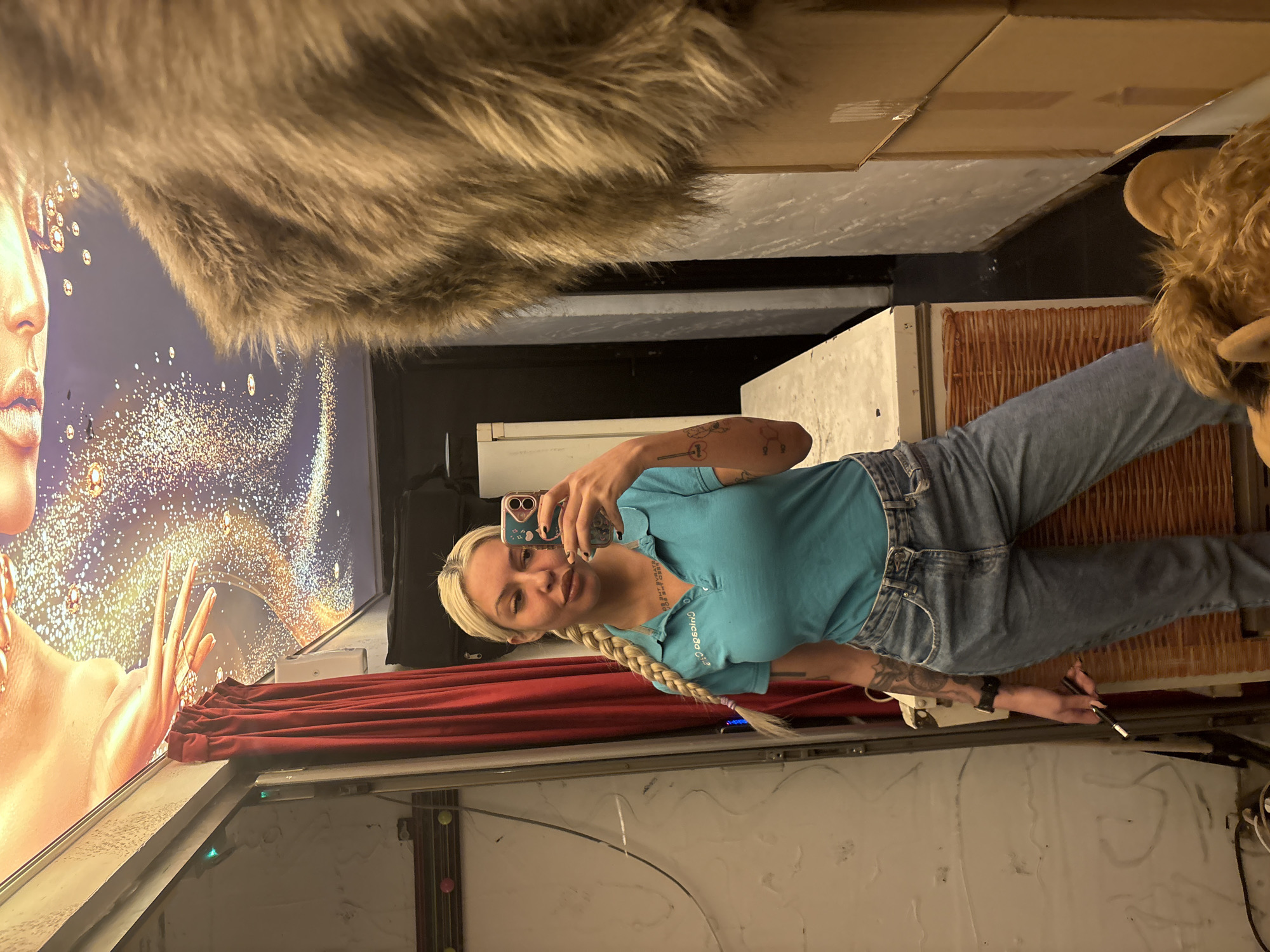A young woman with long blonde hair, styled in a braid, takes a mirror selfie. She is wearing a light blue polo shirt and blue jeans, displaying tattoos on both arms, one depicting a chemical structure and another a key and guitar. She holds a smartphone to capture the image and a thin, dark object, possibly a vape pen, in her right hand.
The setting appears to be an indoor space with various elements. To her left, a large, illuminated poster or display features a close-up of a person's face and hands, adorned with glittering, golden speckles against a dark blue background. Below this display, a red curtain is drawn. Above her head and to the upper left, a fluffy, brown faux fur material is visible, possibly part of the ceiling or a decorative element. To her right, a white refrigerator is partially visible, alongside a brown, textured panel or screen. Cardboard boxes are stacked or placed higher up in the background. The floor is a light, possibly concrete, surface with some dark marks and wires. The text on her polo shirt is mirrored and difficult to decipher, appearing as "da.c odo.cs". The overall lighting is artificial. There are no specific cues in the image to confirm the location is Plovdiv, Bulgaria, other than the provided context.