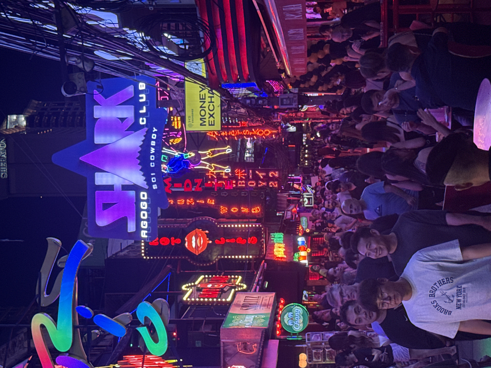 This is a night scene on Soi Cowboy in Bangkok, Thailand, a famous street known for its vibrant nightlife and neon signs. The image captures a bustling street filled with people, illuminated by numerous colorful neon signs. Prominent signs include "SHARK CLUB," "AGOGO," and various bars and establishments with Thai and English text. There are many people walking and standing around, some appear to be patrons of the clubs, while others might be employees. The environment is characterized by the dense overhead wiring typical of Bangkok streets, along with balconies and building facades adorned with lights and advertisements. The overall atmosphere is lively and energetic, typical of a popular entertainment district at night.