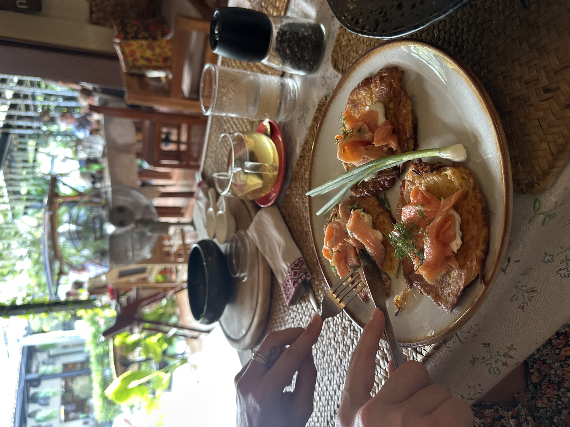 A person is cutting into a meal of latkes with salmon, dill, and green onions with a knife and fork, on a tan-colored plate with a slightly upturned rim, while seated at a table with a woven placemat. To the left of the plate is a small black bowl on a wooden coaster, a white napkin with red trim, and a glass cup on a red saucer. Behind the plate are a clear salt shaker, a pepper grinder, and a glass of water. In the blurred background on the left, there is a glimpse of a fan, vegetation, and outdoor scenery that suggests the location is in Baan Chaweng Noi, Thailand. The person is wearing a floral dress.