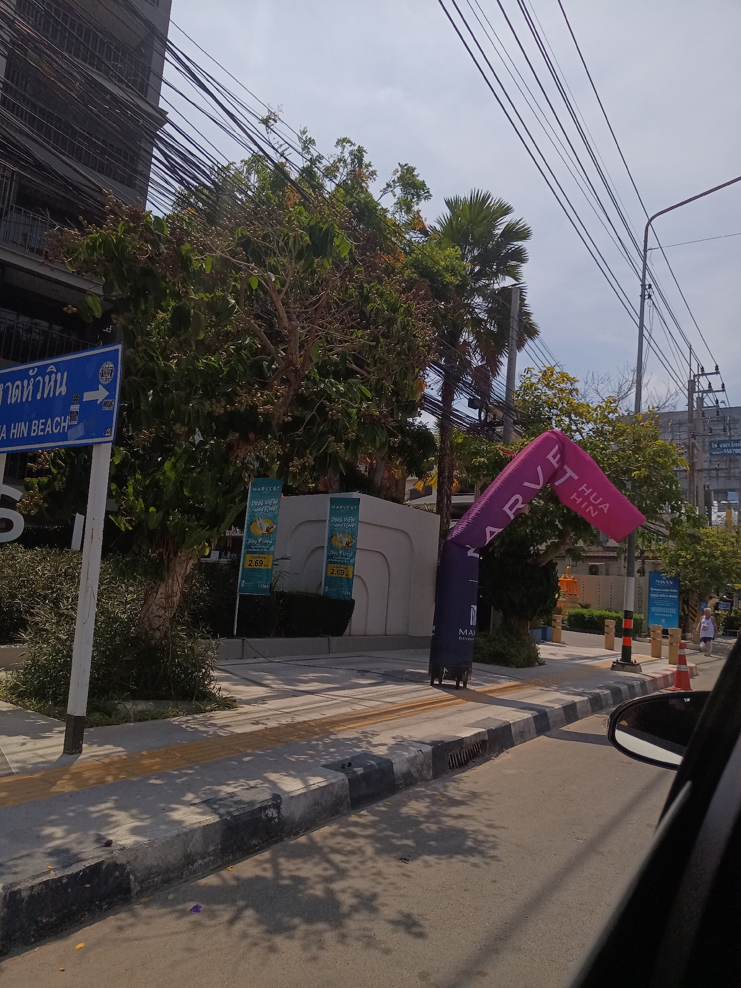 The image captures a street scene in Hua Hin City Municipality, Thailand. A blue sign indicating "Hua Hin Beach" stands on the left. In the background, there is a building obscured by dense trees and a web of overhead power lines. A purple and black inflatable archway with "MARVEST HUA HIN" printed on it marks the entrance to a property. Promotional signs for "MARVEST" advertise pool view units. Orange traffic cones line the sidewalk. A glimpse of a car's side mirror suggests the photo was taken from inside a vehicle. A person is walking on the sidewalk in the background.
