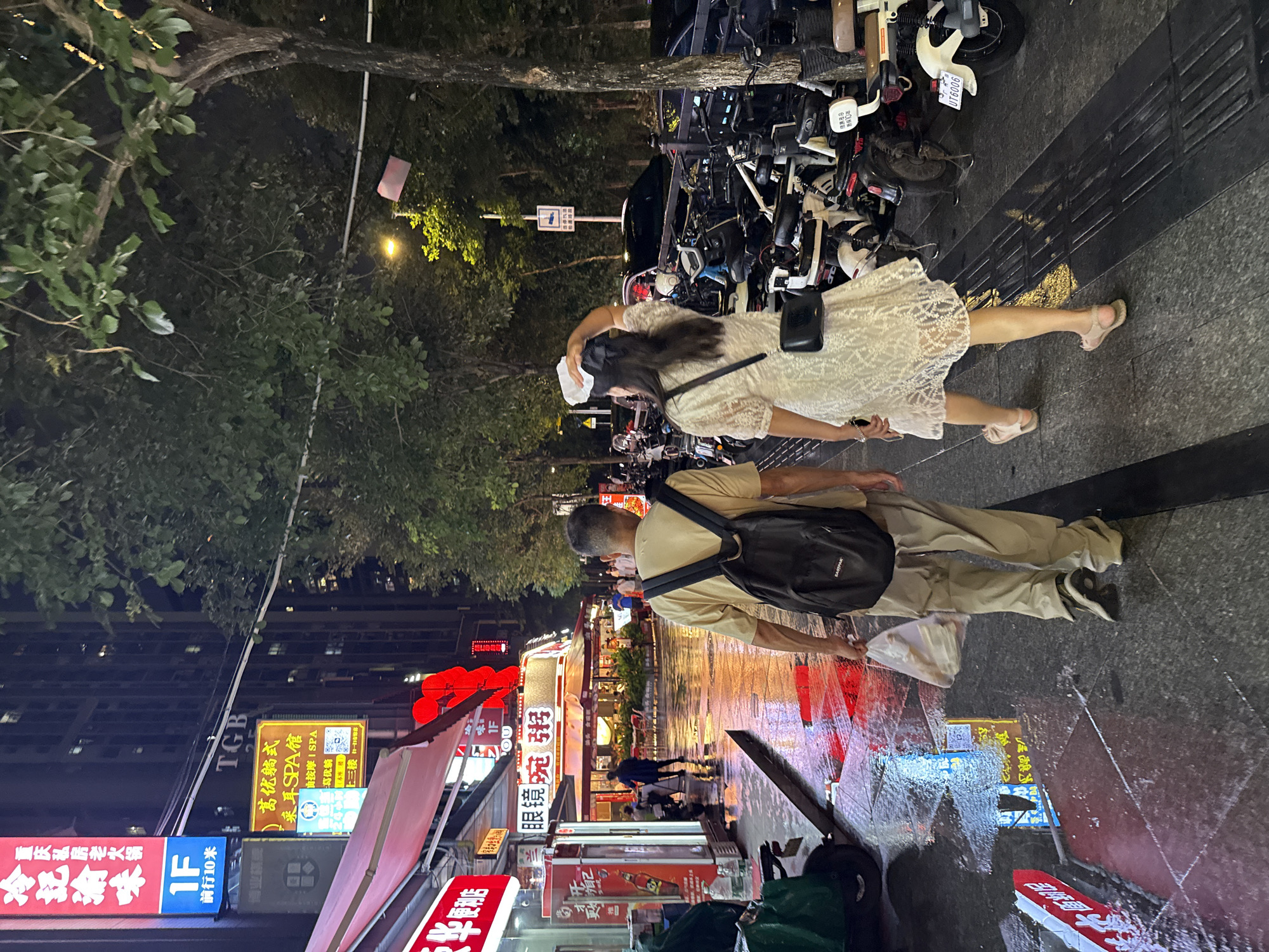A night scene unfolds on a wet city street in Haizhu District, China, illuminated by the vibrant glow of neon signs reflecting off the slick pavement. Two people are walking away from the camera, their figures silhouetted against the illuminated storefronts. In the foreground, a man in a tan shirt and light-colored pants carries a black backpack, his hand holding a white plastic bag. Beside him, a woman in a white lace dress walks, her dark hair cascading down her back.
The street is lined with shops, indicated by various signs displaying Chinese characters, some of which are partially legible: "重庆私房老火锅" (Chongqing Private Hot Pot), "采耳SPA馆" (Ear Cleaning SPA), "博业棋牌" (Boye Chess and Cards), and "眼镜碗粥" (Glasses Bowl Porridge). One sign also shows "TGB" and "UT6006". The presence of many parked motorcycles suggests this is a busy commercial area. The wet street and reflections indicate recent rain or ongoing drizzle, contributing to the atmospheric ambiance of the nighttime urban setting. The overall mood is one of casual evening activity in a lively commercial district.