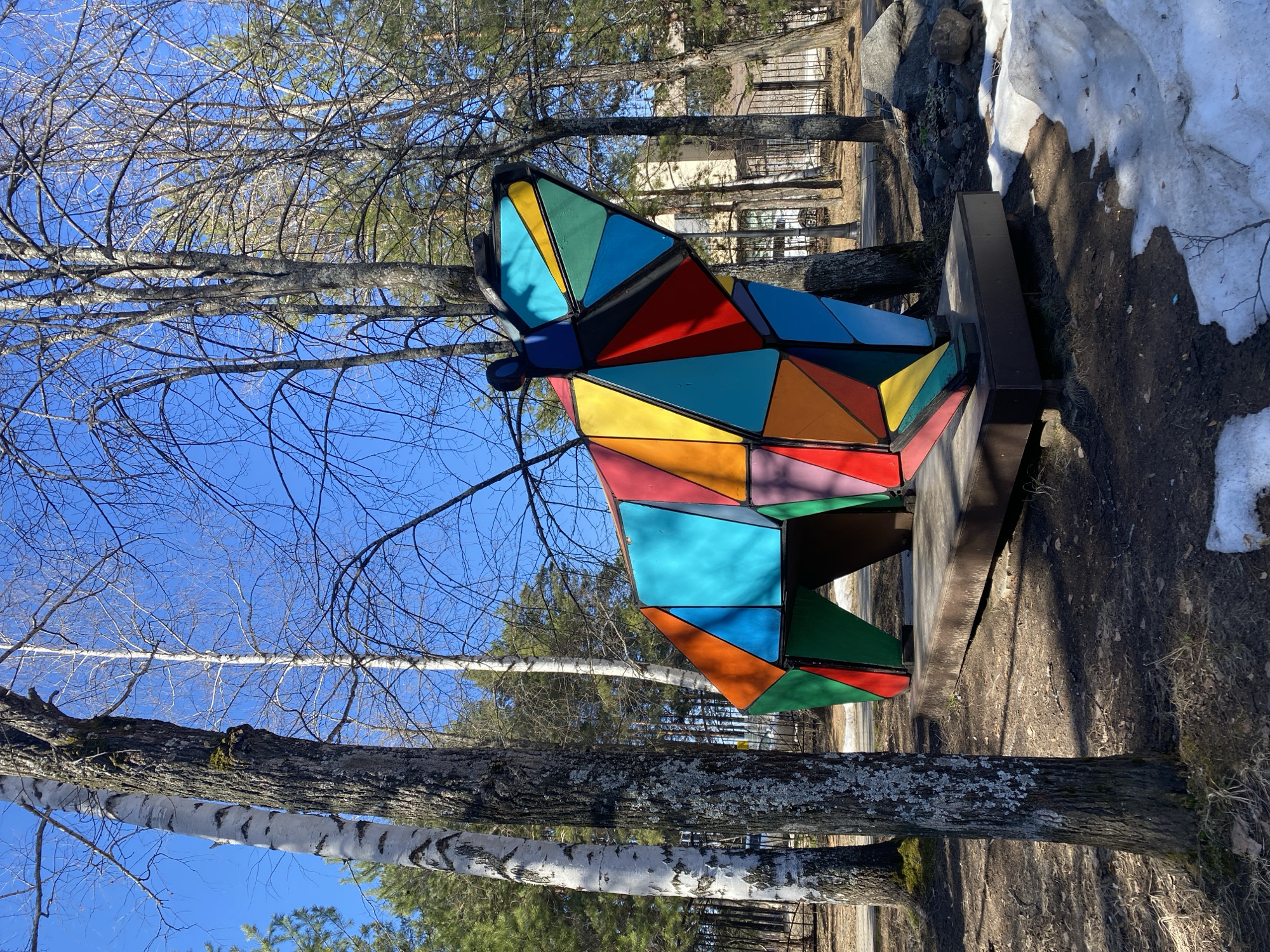 The image captures a sunny outdoor scene in what appears to be a park setting. The main subject is a large, geometric butterfly sculpture made of colorful triangular segments. The sculpture is mounted on a dark platform, which is positioned near a tree. The background includes bare trees with white trunks and branches reaching toward a clear blue sky. Patches of snow can be seen in the background. This scene is specifically located in the city of Chaikovsky, Russia.